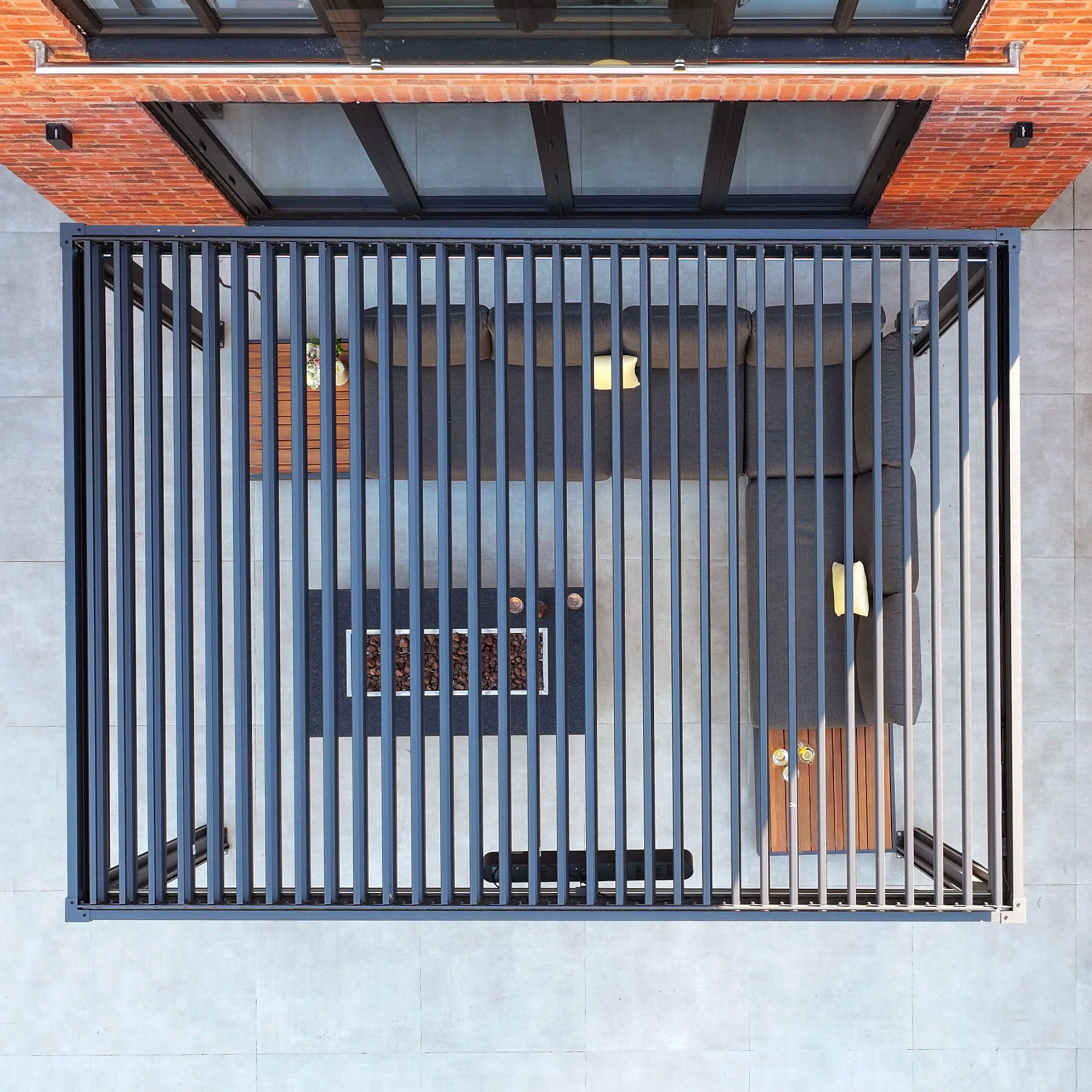 Aerial view of a sleek patio with gray sofas and a rectangular fire table beneath PergoSTET Plus 3m x 4m Rectangular Pergola in Grey, featuring vertical slats and LED lighting against a brick wall.