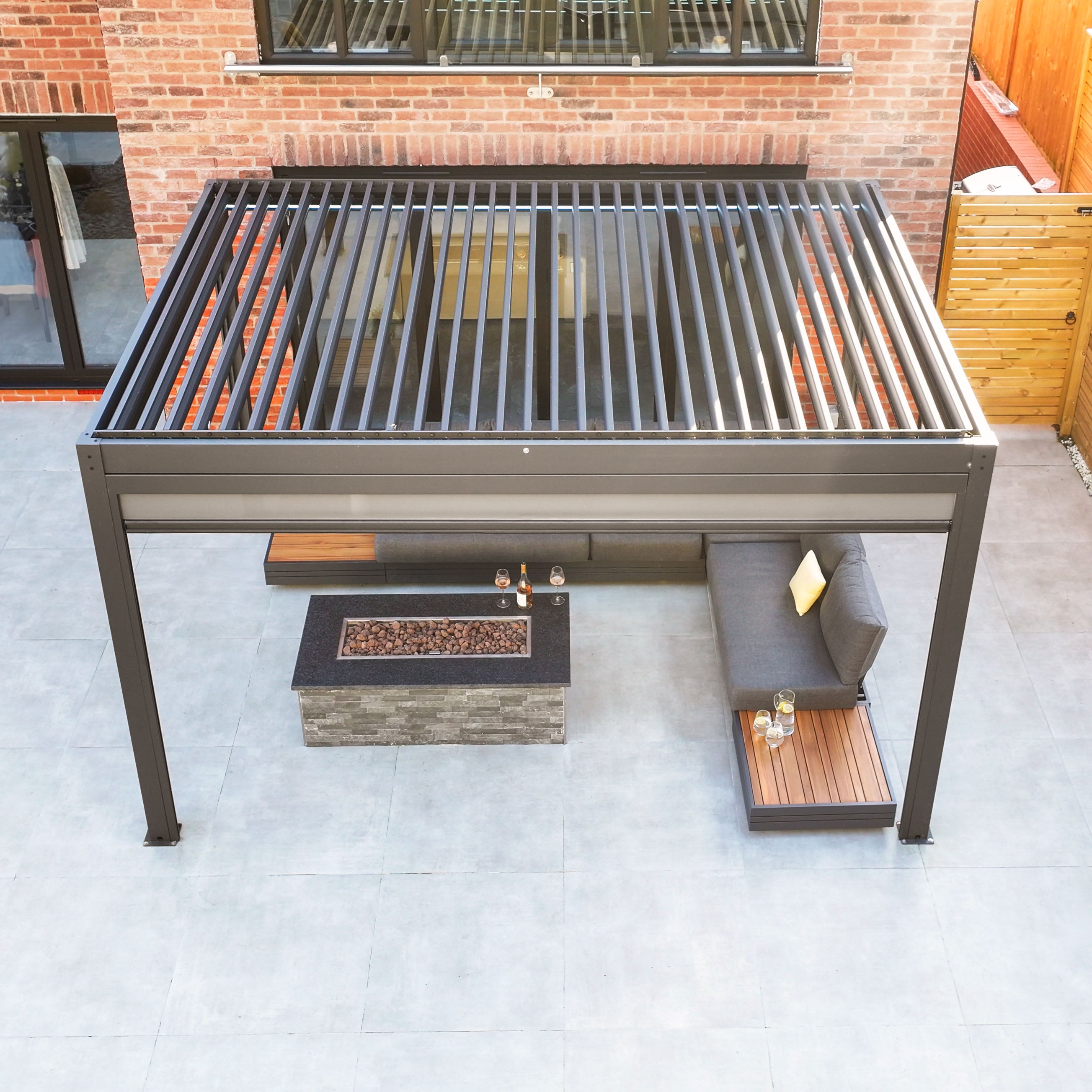 The PergoSTET Plus 3m x 4m pergola, featuring adjustable slats, textilene blinds, and LED lighting in grey, enhances the patio seating area with a fire pit besides the charming brick building.