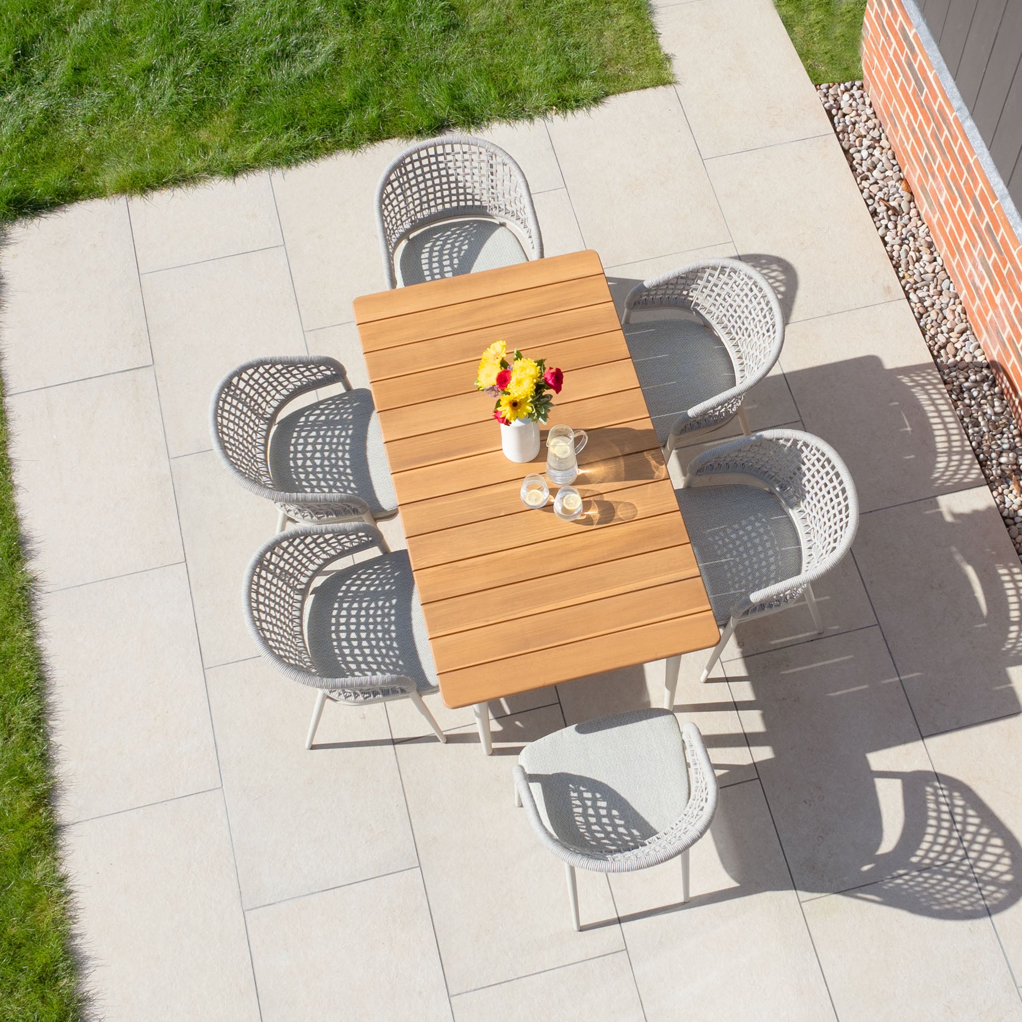 Aerial view of the patio featuring the Kalama 6 Seat Rectangular Dining Set with Teak Table in Latte, showcasing a teak table, six chairs, and a colorful flower vase basking in sunlight.