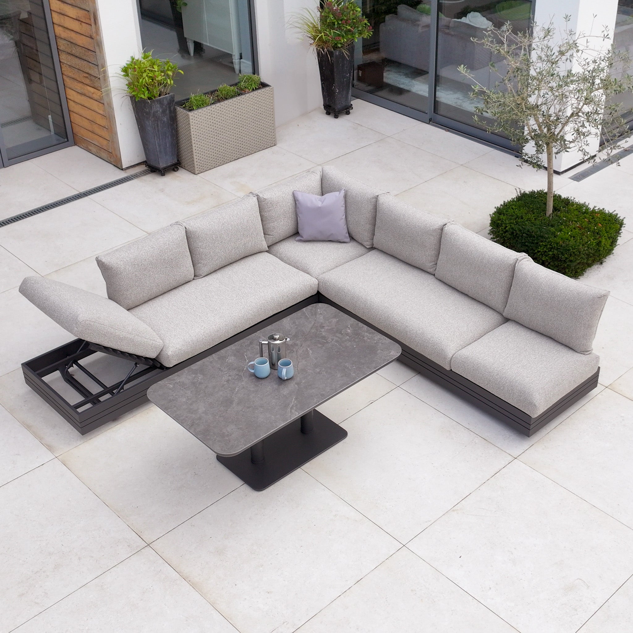 The Savona Luxury Outdoor Large Corner Sofa Set with Altura Rising Table in Truffle creates a modern patio look, featuring a spacious sectional and stylish rising table on tiled flooring.