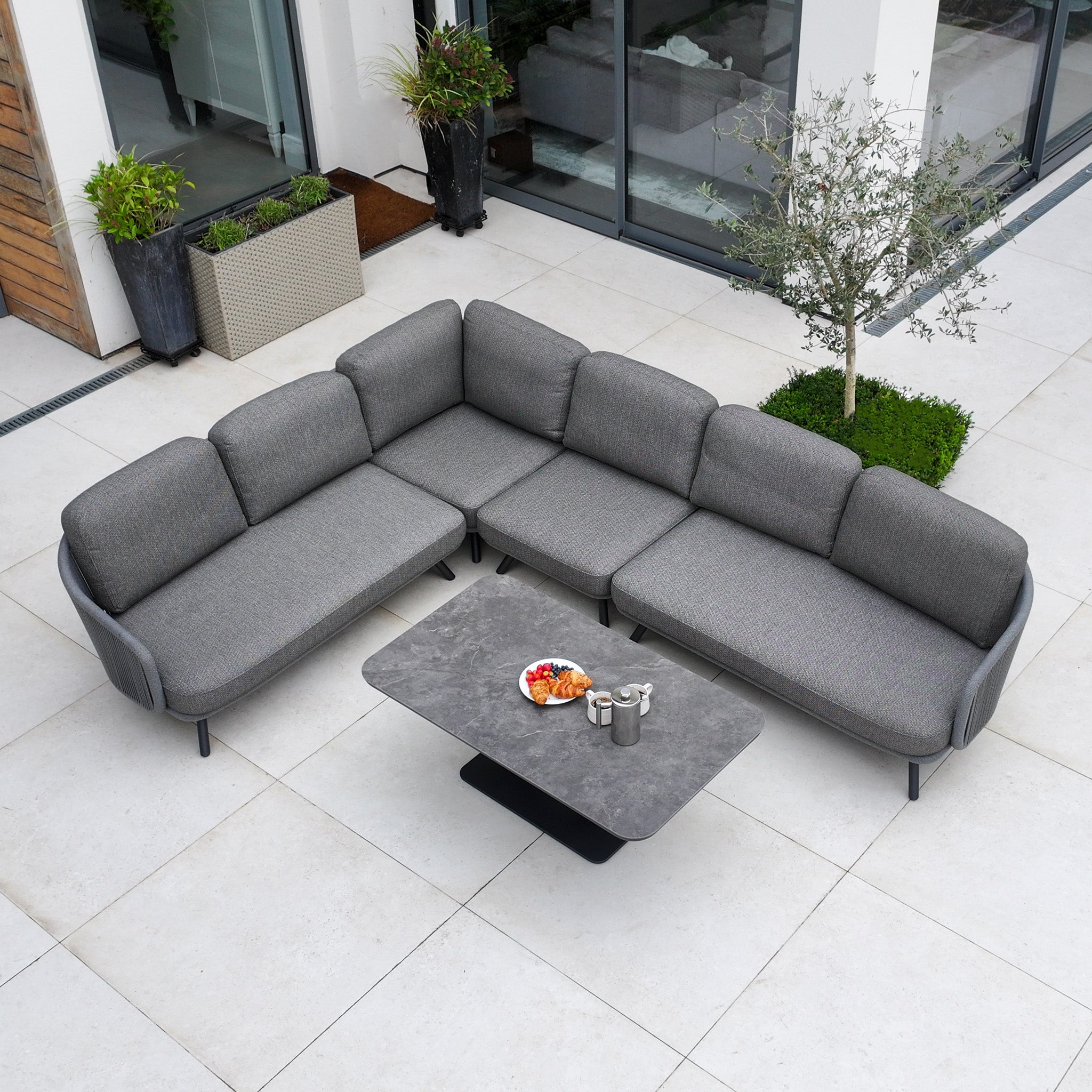 The Dekota Large Corner Sofa Dining Set with Altura Rising Table in Charcoal is displayed on a modern patio next to a building and lush plants, complete with snacks arranged for outdoor entertaining.