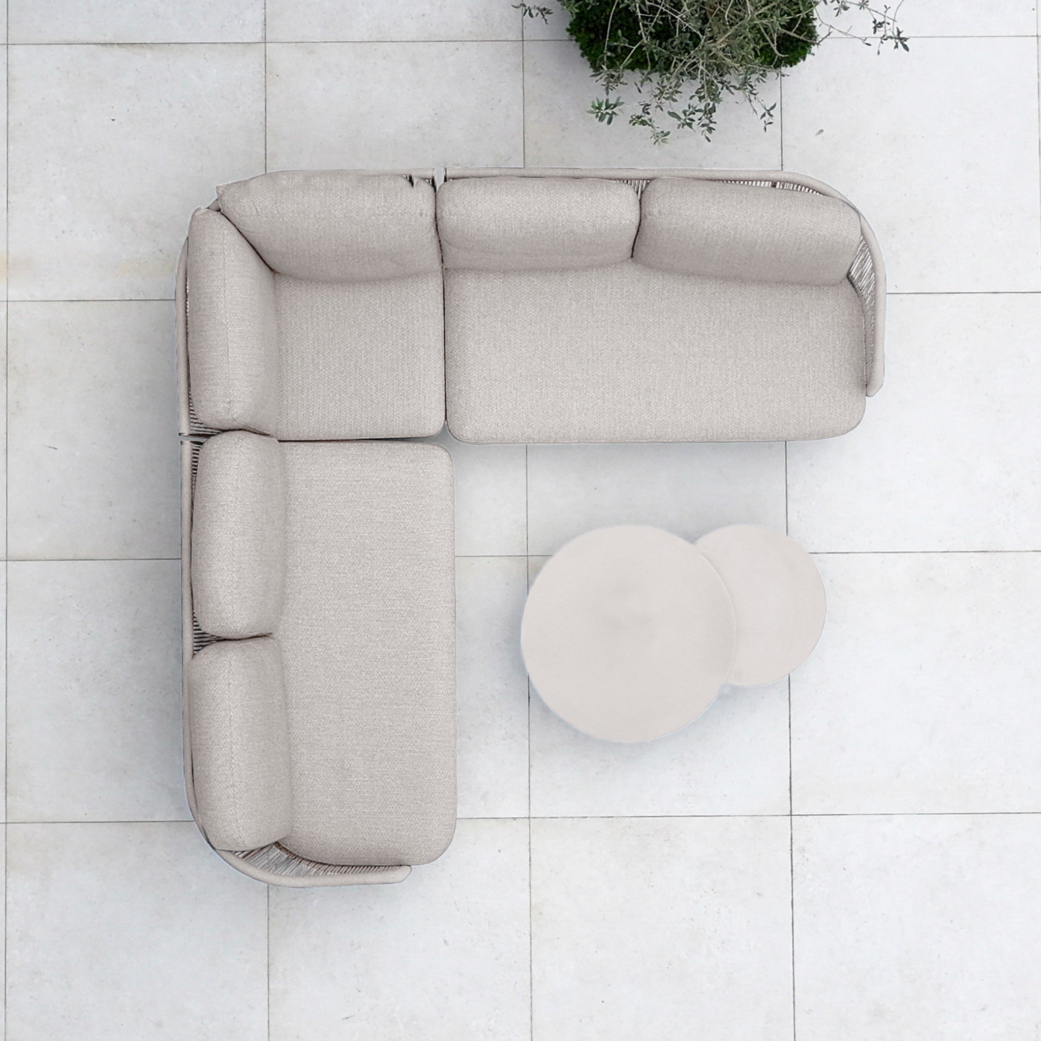 Top view of the Dekota Corner Sofa Set in Fawn—an L-shaped beige outdoor piece with hydrophobic rope weave—paired with two round nesting tables on a tiled floor beside a green plant.