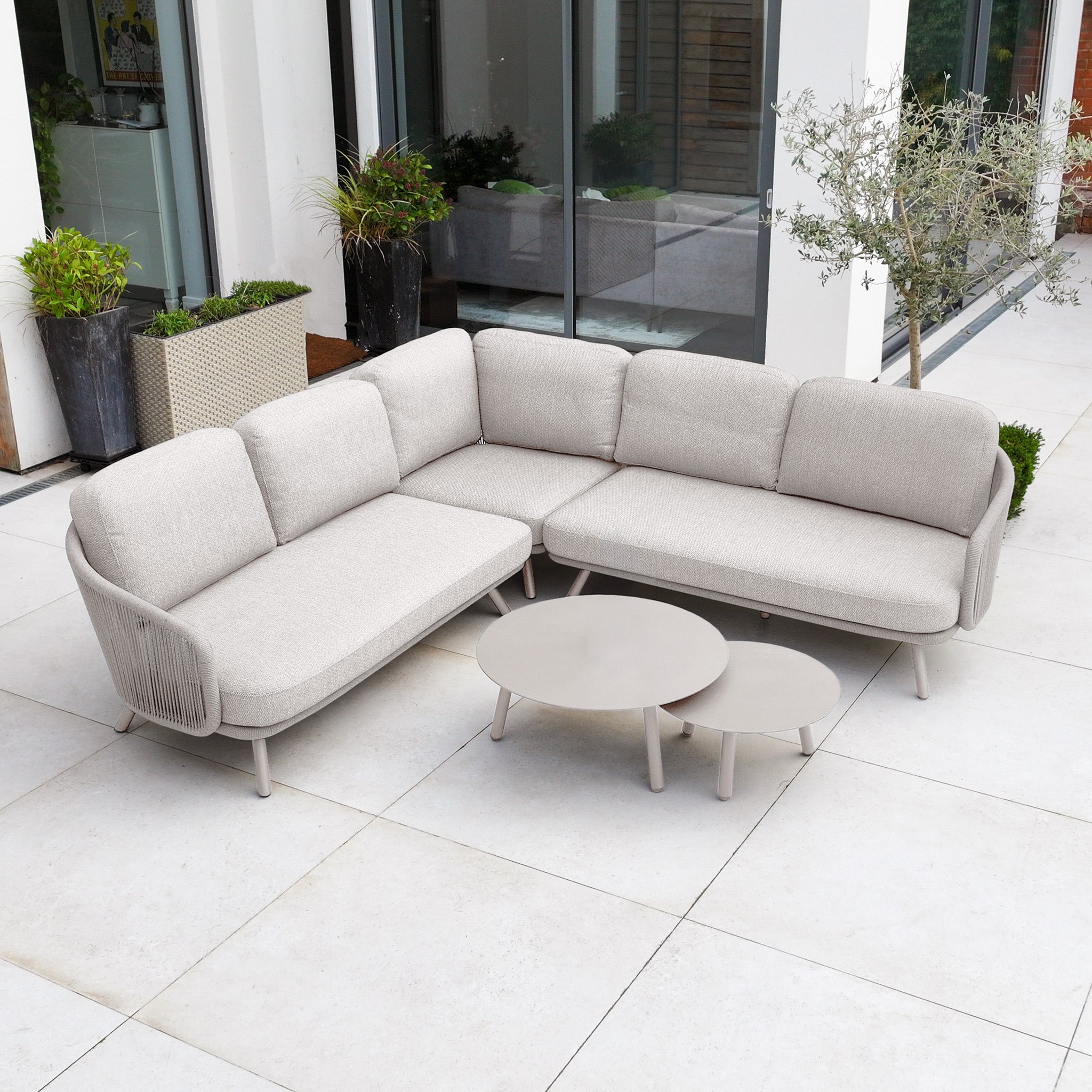 Dekota Corner Sofa Set in Fawn with hydrophobic rope weave, plus two round coffee tables on a tiled patio by large windows and potted plants—ideal for stylish outdoor furniture arrangements.