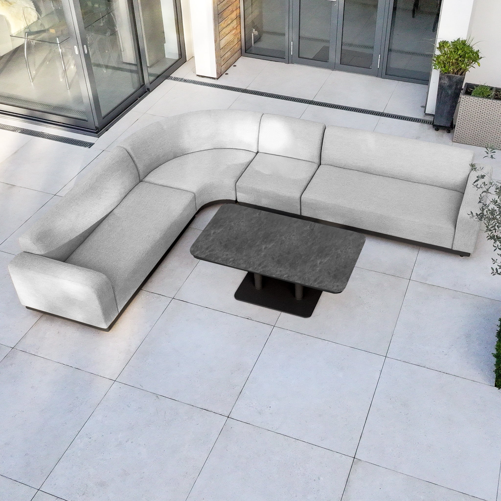 Modern outdoor patio with a Luna Outdoor Fabric Large Curved Corner Sofa Set in Oyster Grey and an Altura Rising Table, arranged on large stone tiles, featuring durable all-weather fabric for lasting style.