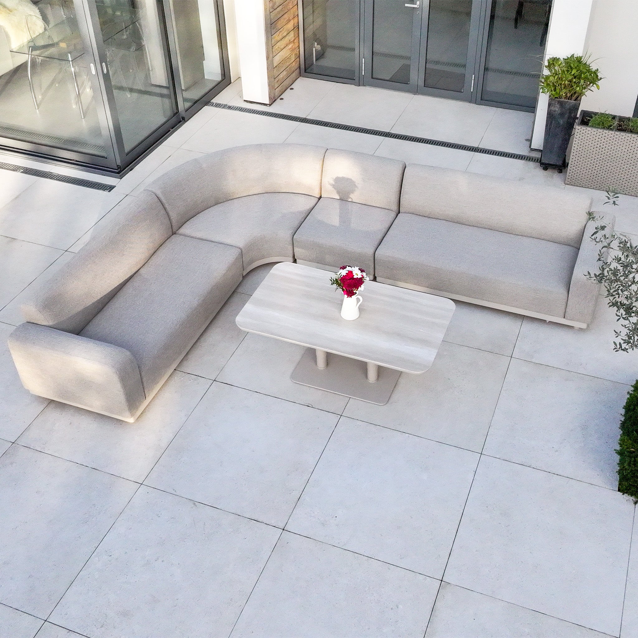 The Luna Outdoor Fabric Large Curved Corner Sofa Set in Fawn with quick-dry cushions is paired with an Altura Rising Table on a tiled patio beside a vase of flowers.