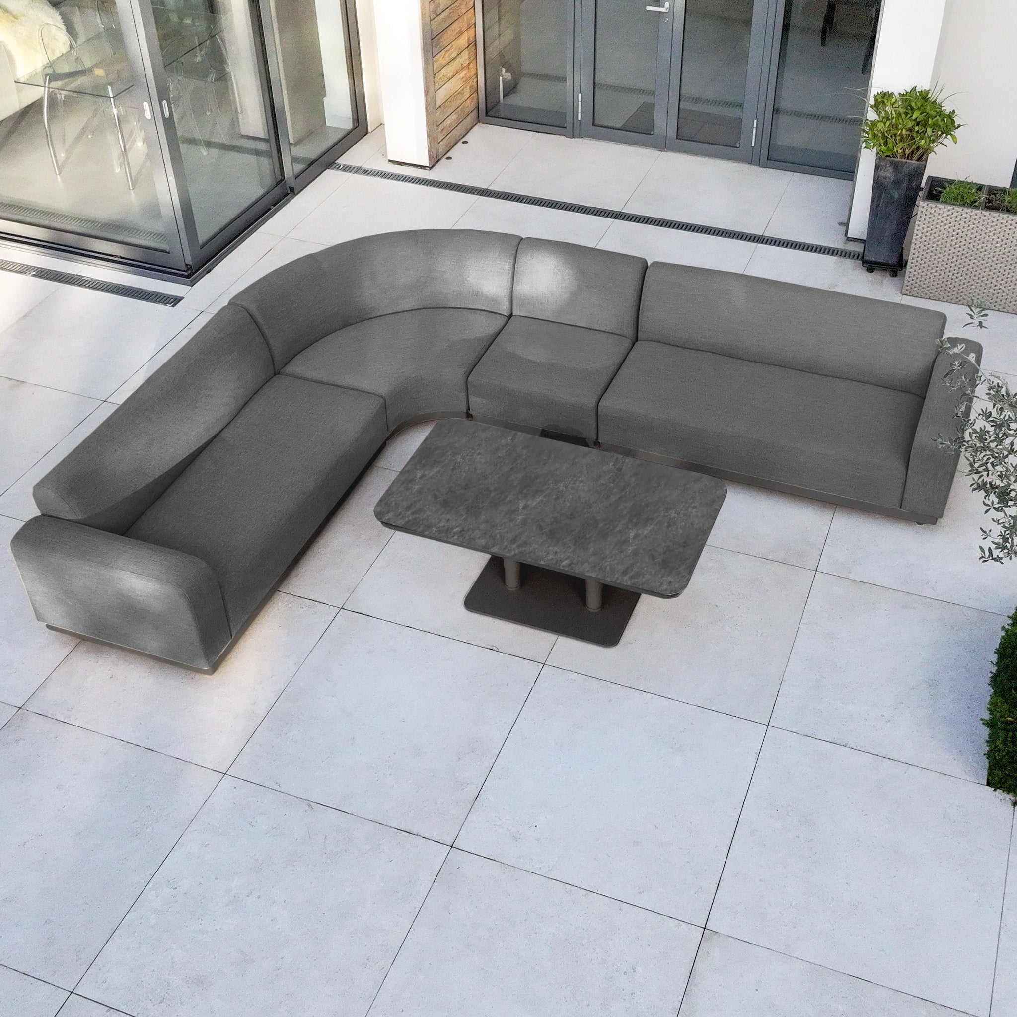 A Luna Outdoor Fabric Large Curved Corner Sofa Set with Altura Rising Table in Grey sits on a tiled patio by glass doors and plants, offering stylish comfort and all-weather durability for outdoor living.