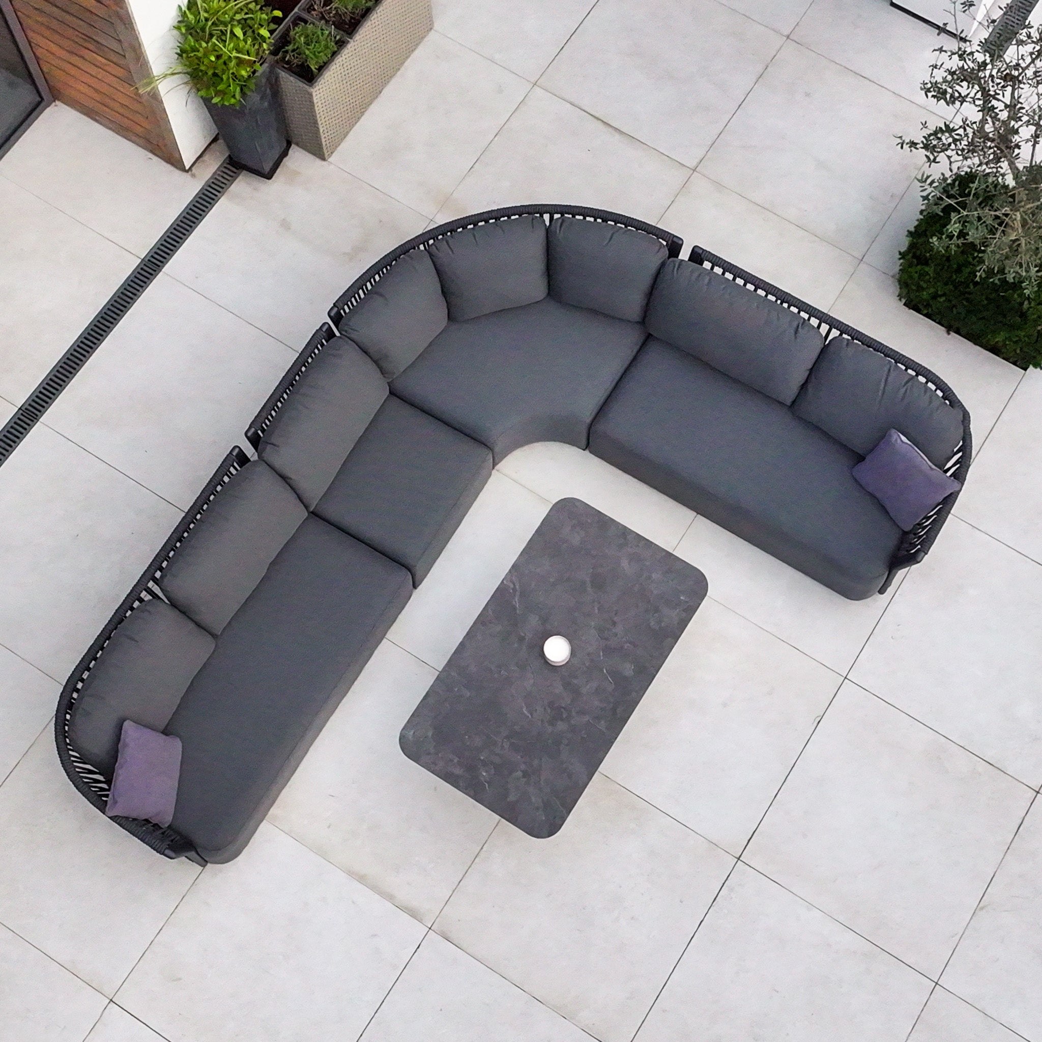 The Pello Deluxe Curved Sofa Set with Altura Rising Table in Grey is a stylish, comfortable outdoor L-shaped sofa set with two purple pillows and a rectangular rising table, perfect for enhancing any garden patio.
