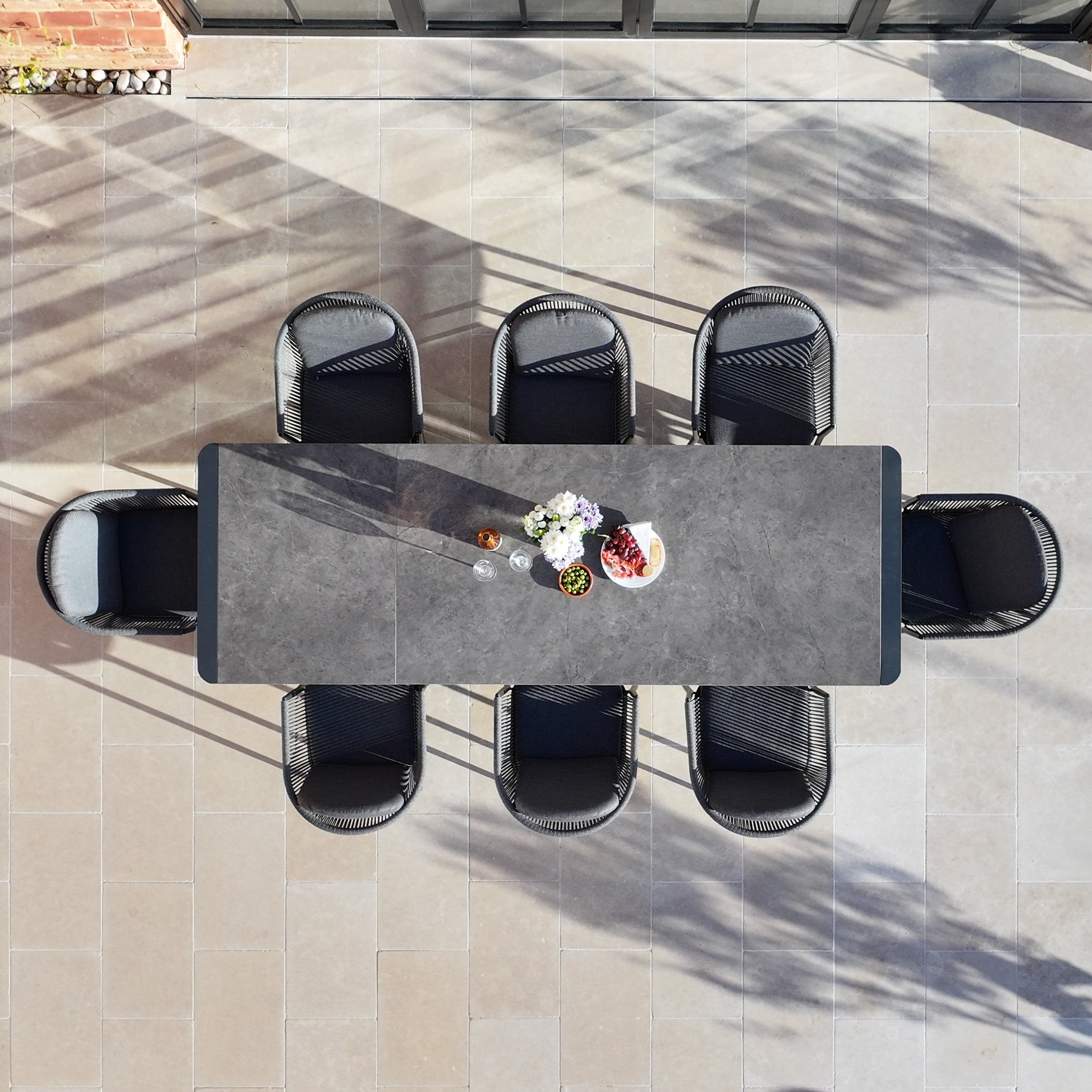 The Ravona 8 Seat Rope Extending Dining Set (207-277cm) in Charcoal, shown with eight chairs and decor on a tiled patio, is perfect for stylish outdoor gatherings.