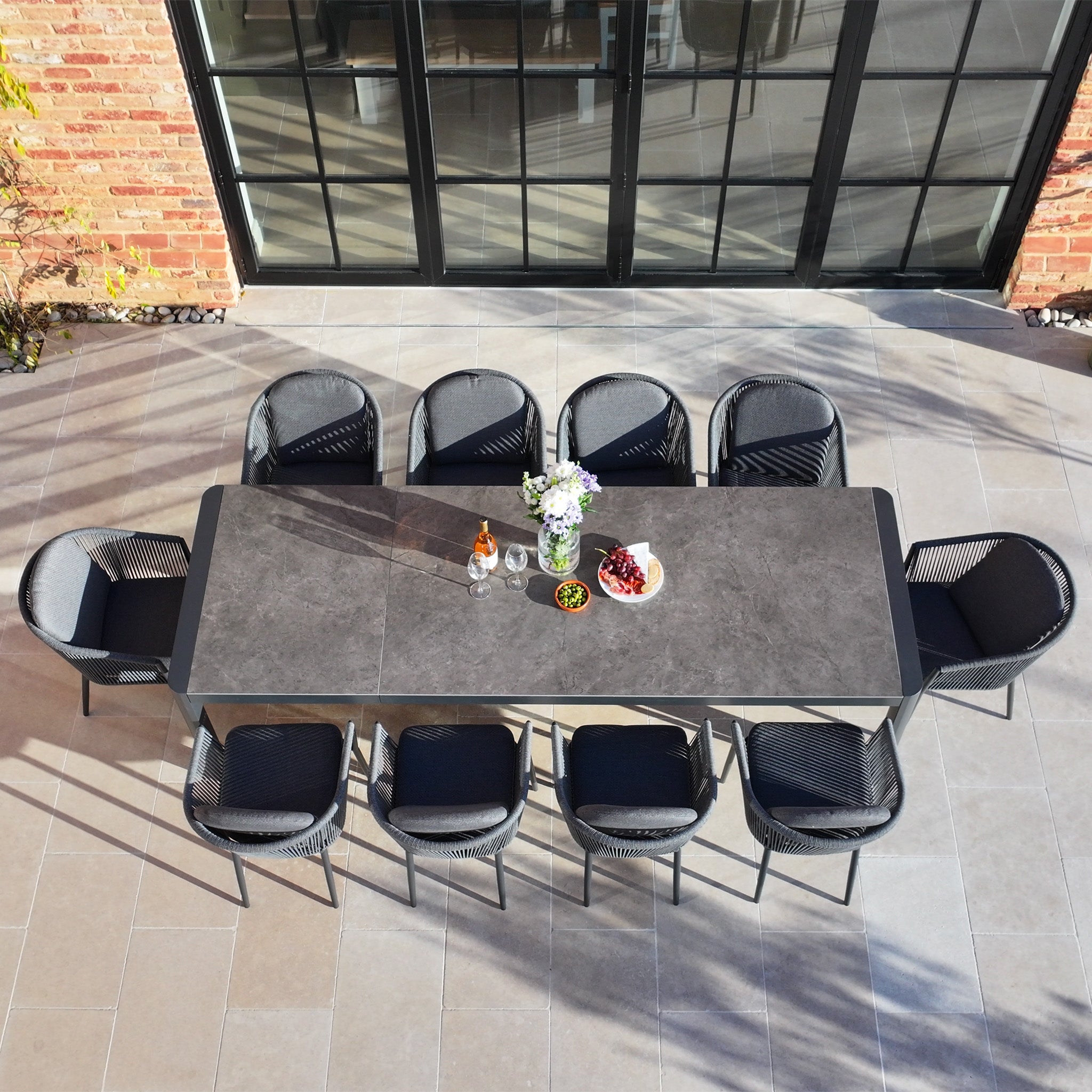 The Ravona 10 Seat Rope Extending Dining Set (207-277cm) in Charcoal, arranged with drinks, flowers, and fruit on a tiled patio, is perfect for stylish outdoor gatherings.