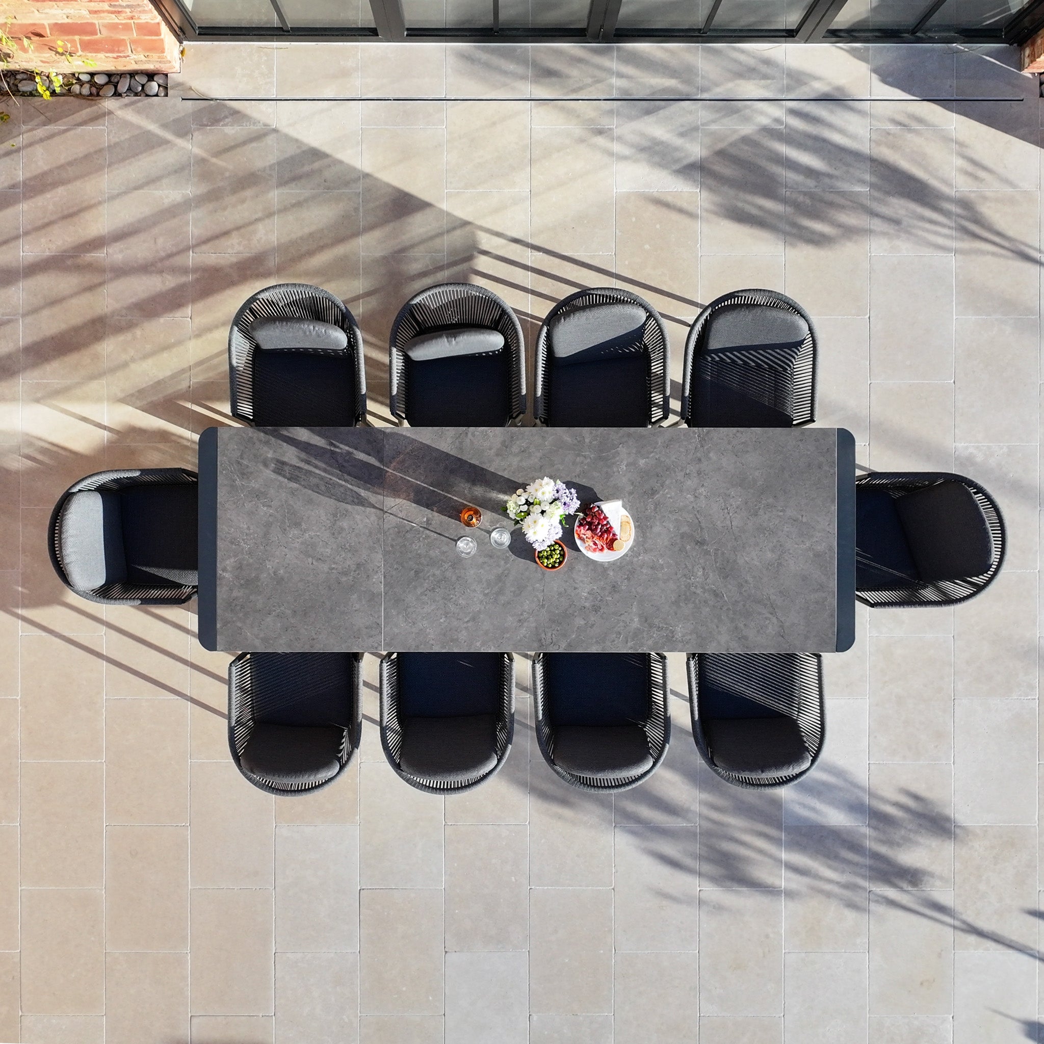 The Ravona 10 Seat Rope Extending Dining Set (207-277cm) in Charcoal, rectangular and set for dining, is shown from above with eight chairs on a tiled patio.