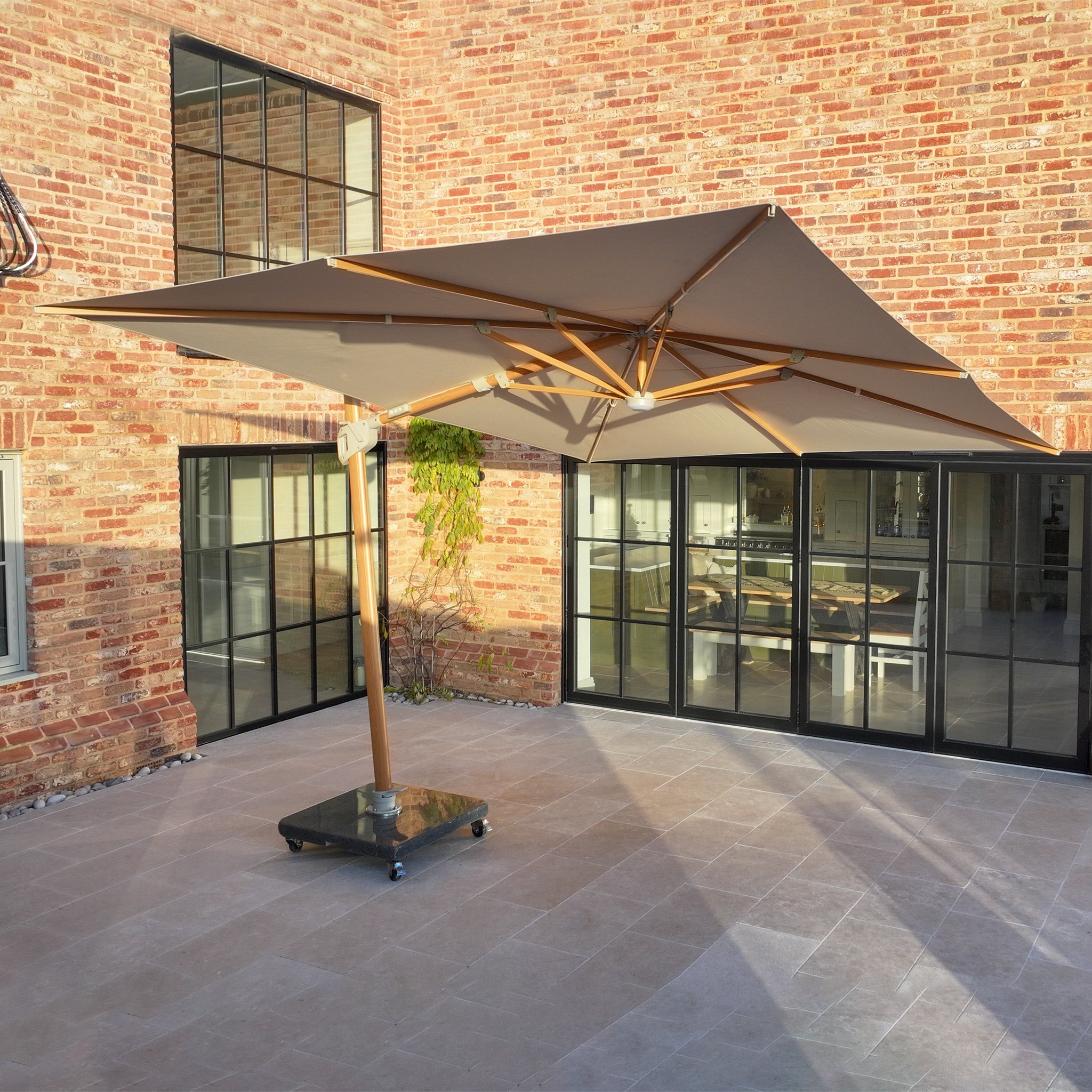 The Volos 3m x 4m Rectangular Cantilever Parasol in Wheat with LED lighting and a 120kg granite base stands on wheels in a sunny courtyard with brick walls and glass doors.