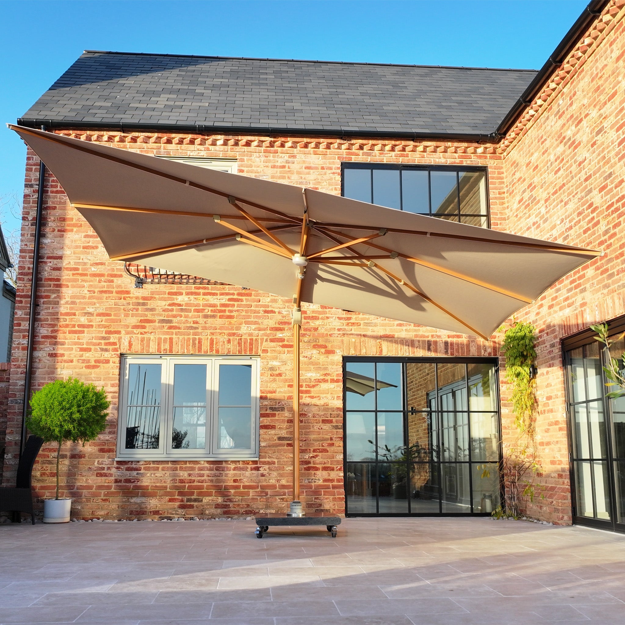 The Volos 3m x 4m rectangular cantilever parasol in wheat with wood effect, LED lighting, and a 120kg granite base stands on a tiled terrace outside a modern brick house with large windows.