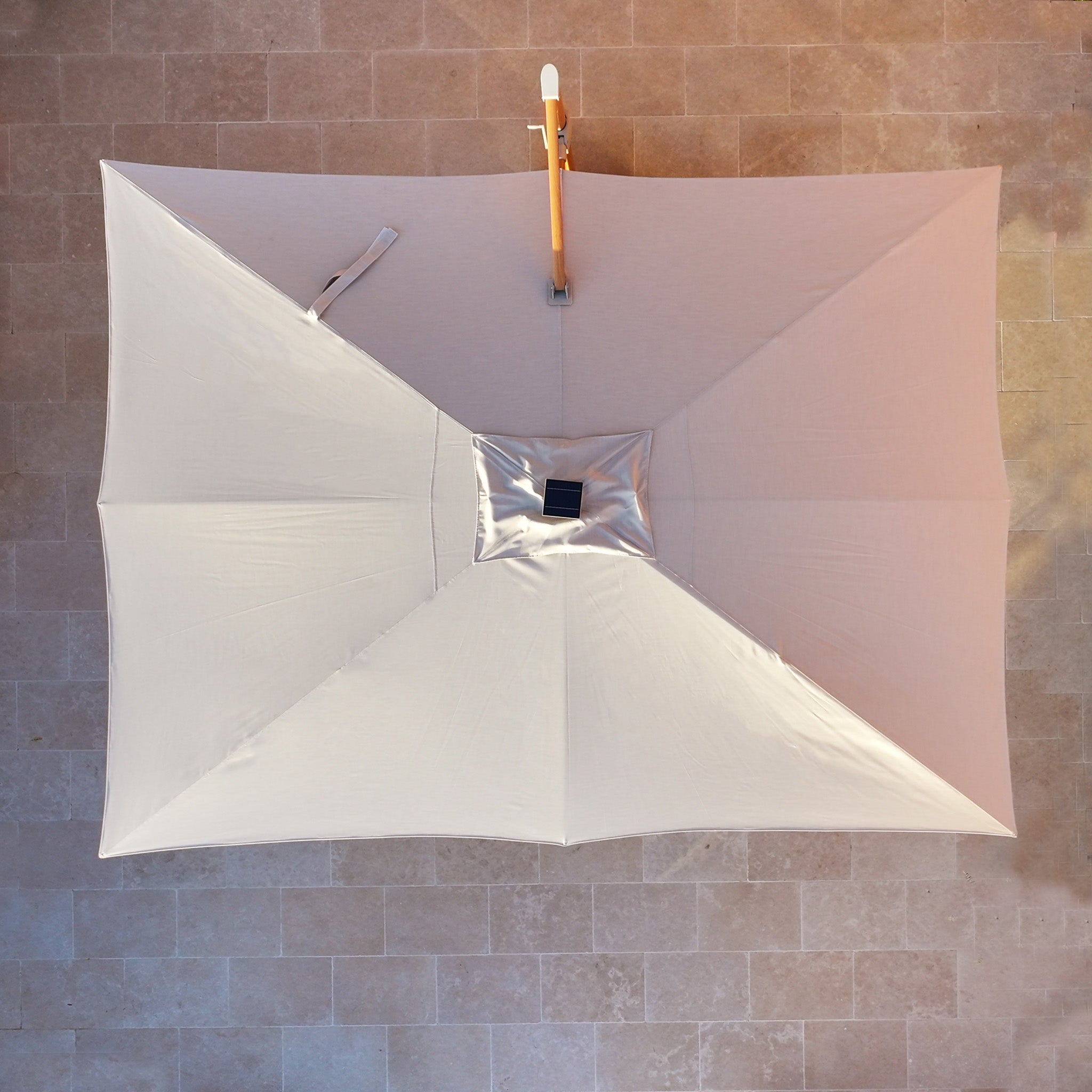 The Volos 3m x 4m rectangular cantilever parasol in Wheat features a UV-resistant canopy, LED lighting, and a sturdy 120kg granite base with a wood effect, casting shade over stone tiles.