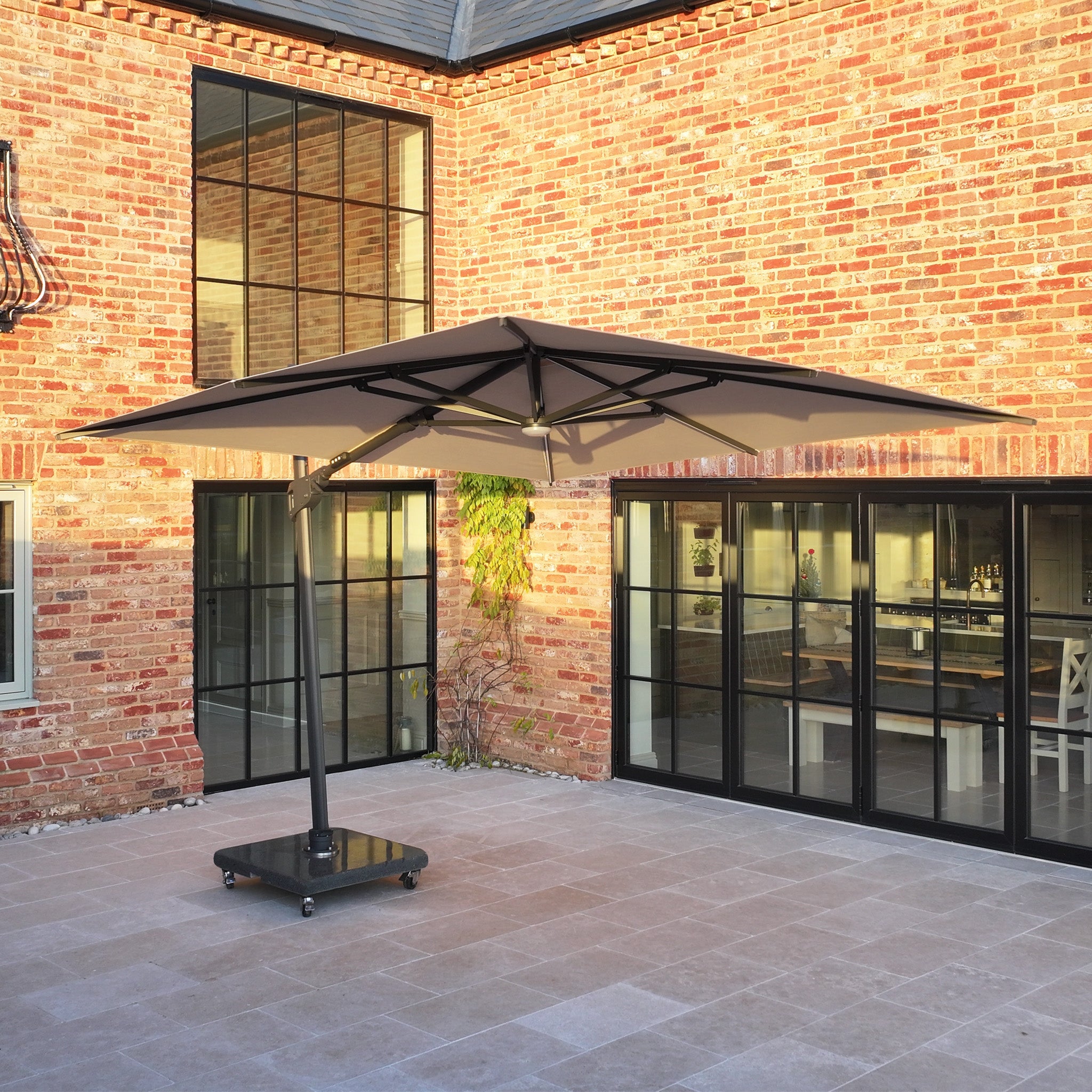 The Volos 3m x 3m Square Cantilever Parasol with LED Lighting and a 120kg granite base in Wheat shades a tiled courtyard beside a modern brick house with large glass doors.