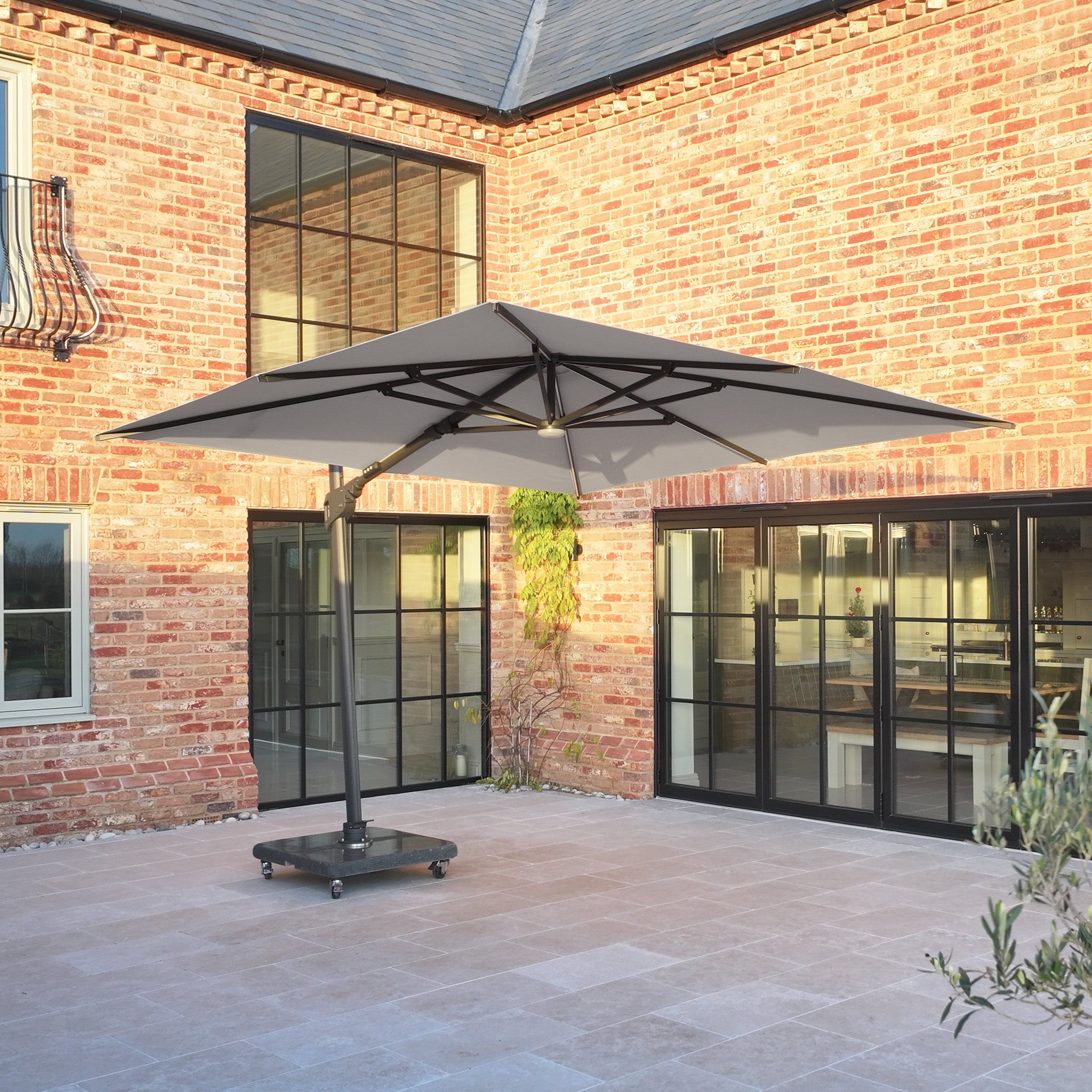 The Volos 3m x 4m Rectangular Cantilever Parasol with LED lighting and a 120kg granite base in Slate stands on wheels in a paved courtyard by a brick house with glass doors.
