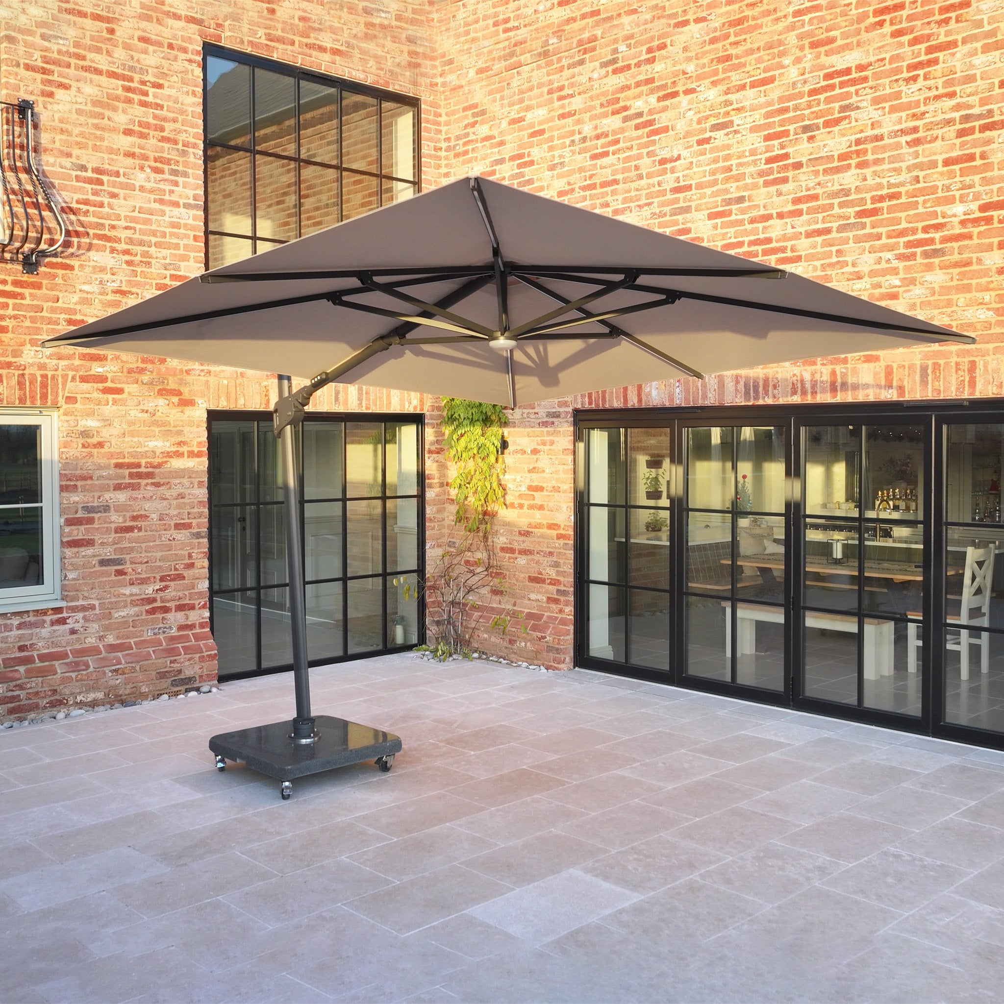 The Volos 3m x 4m Aluminium Cantilever Parasol with LED Lighting and 120kg Granite Base in Wheat stands in a sunlit courtyard with brick walls and large glass doors.