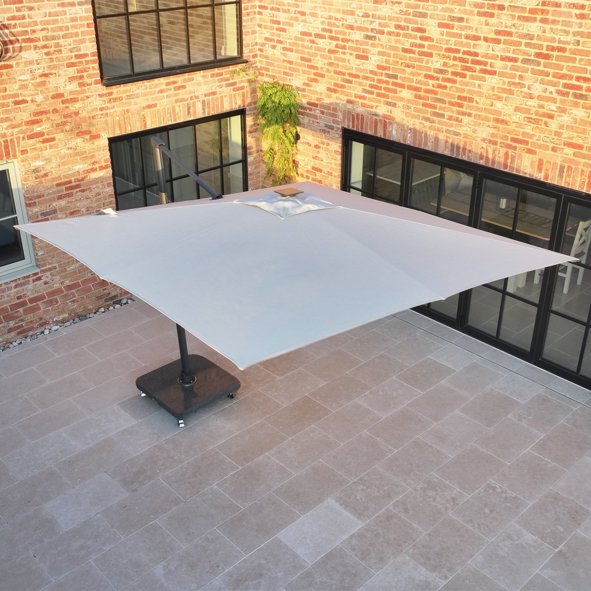 The Volos 3m x 3m Square Cantilever Parasol with LED Lighting and a 120kg granite base in Wheat shades the tiled courtyard beside a brick building with numerous windows and glass doors.