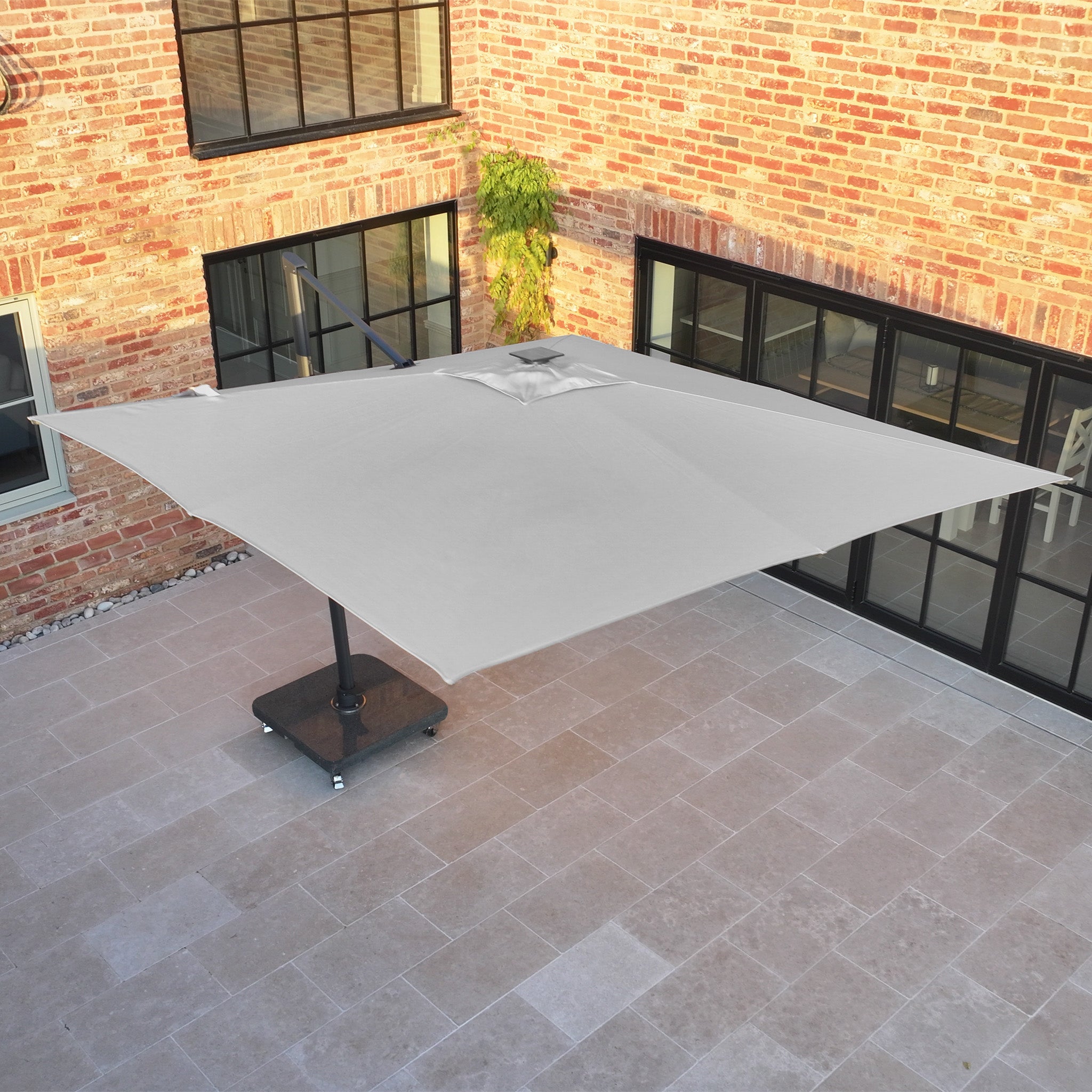 The Volos 3m x 4m Rectangular Cantilever Parasol with LED lighting and a 120kg granite base in Slate offers UV-protected shade on a stone-tiled courtyard beside a brick building.