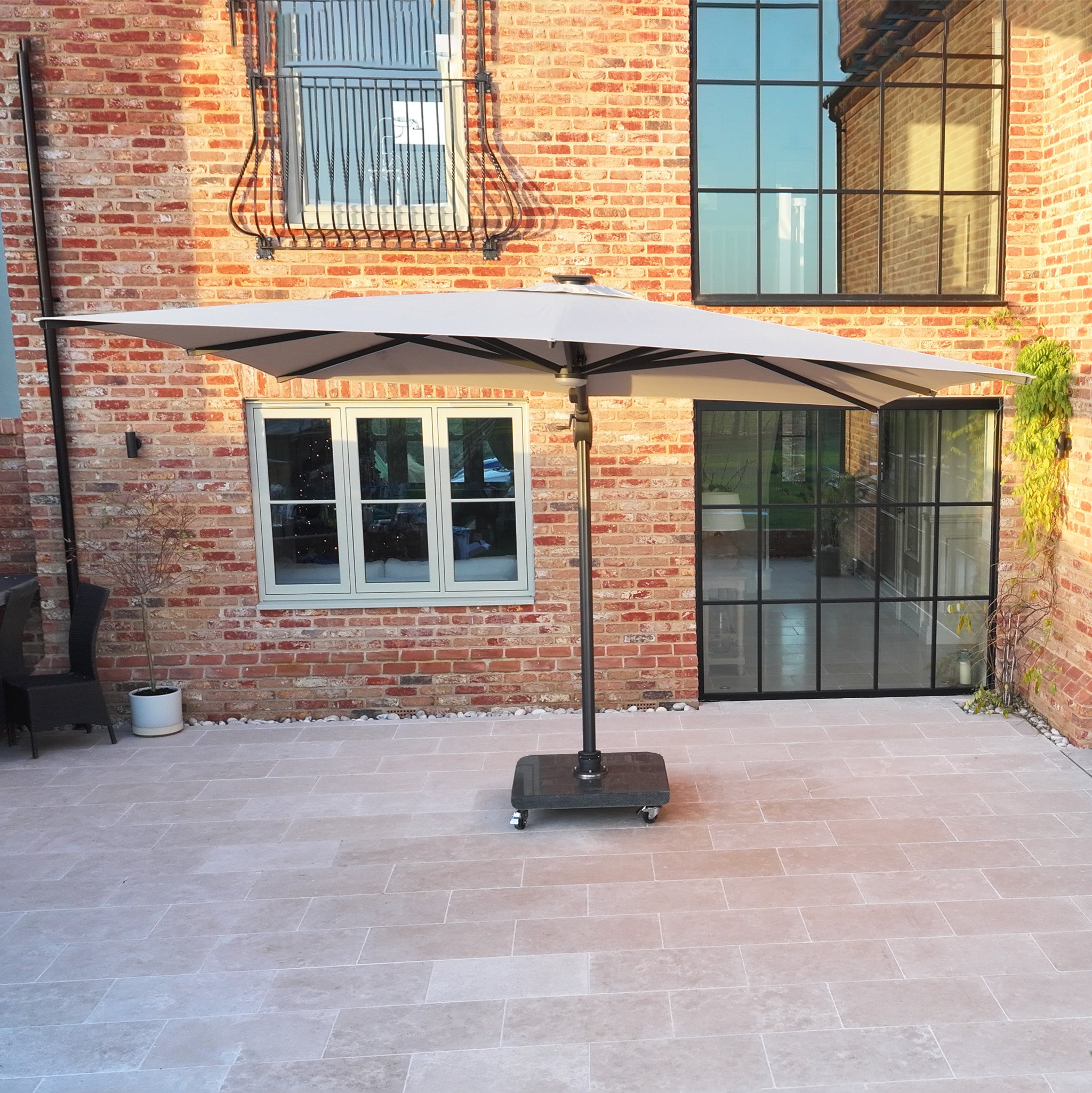 The Volos 3m x 4m Aluminium Cantilever Parasol in Wheat with LED lighting and a 120kg granite base stands on a tiled terrace before a modern brick house with glass doors.