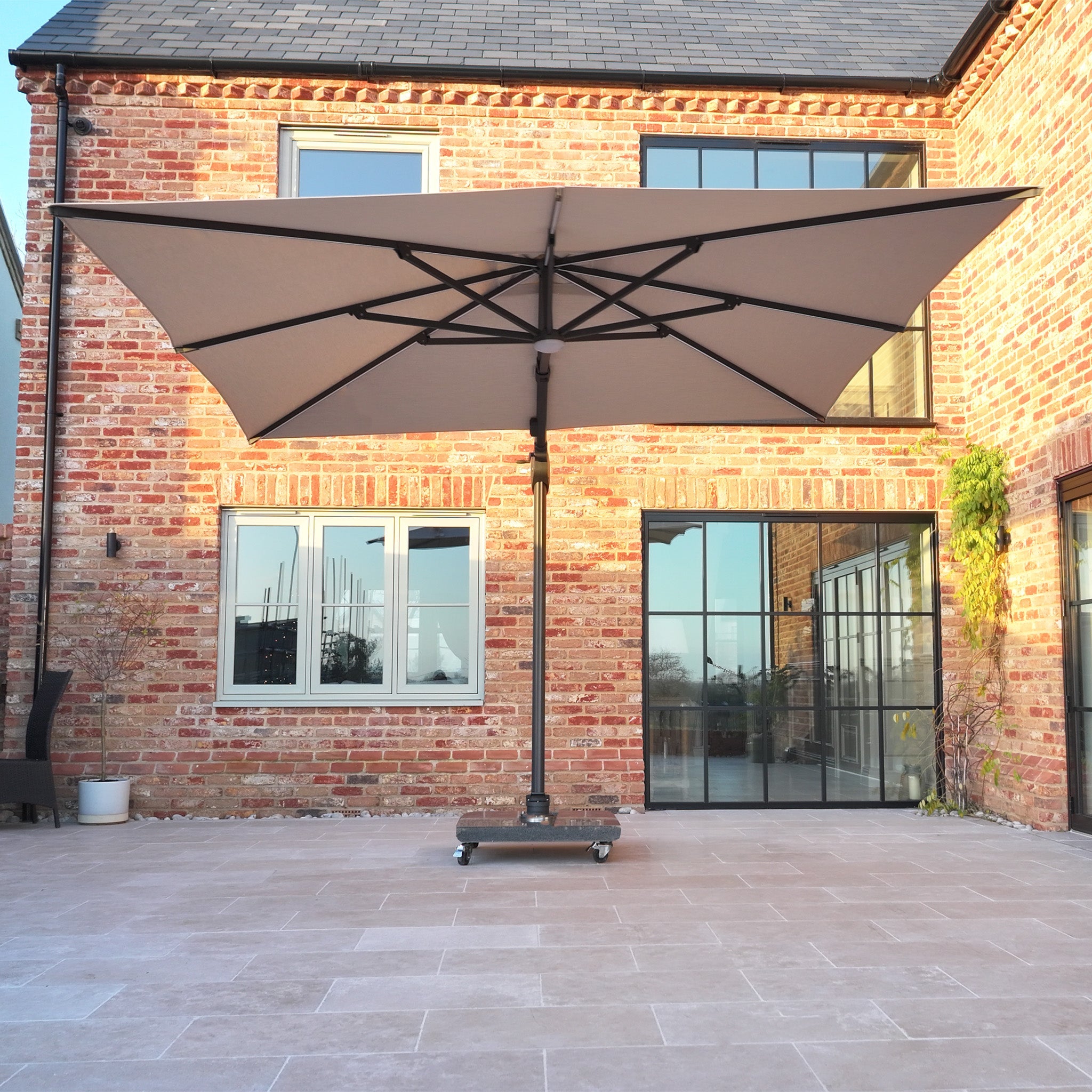 The Volos 3m x 4m Aluminium Cantilever Parasol with LED Lighting and a 120kg granite base in Wheat stands on a tiled patio before a modern brick house with glass doors and windows.
