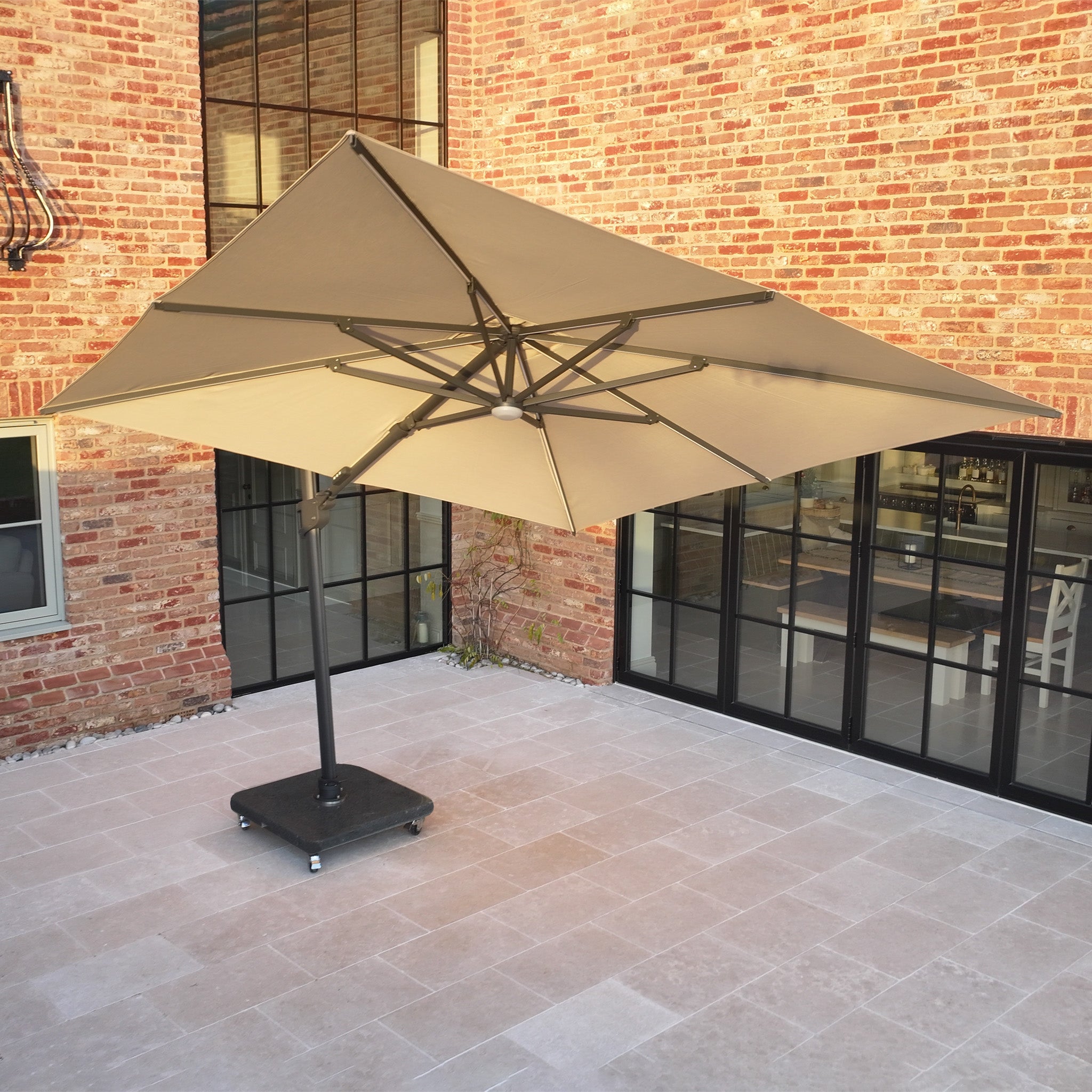 The Volos 3m x 4m Aluminium Cantilever Parasol in Wheat, with LED lighting powered by solar charging and a 120kg granite base, stands on a tiled courtyard beside a brick building with large windows.