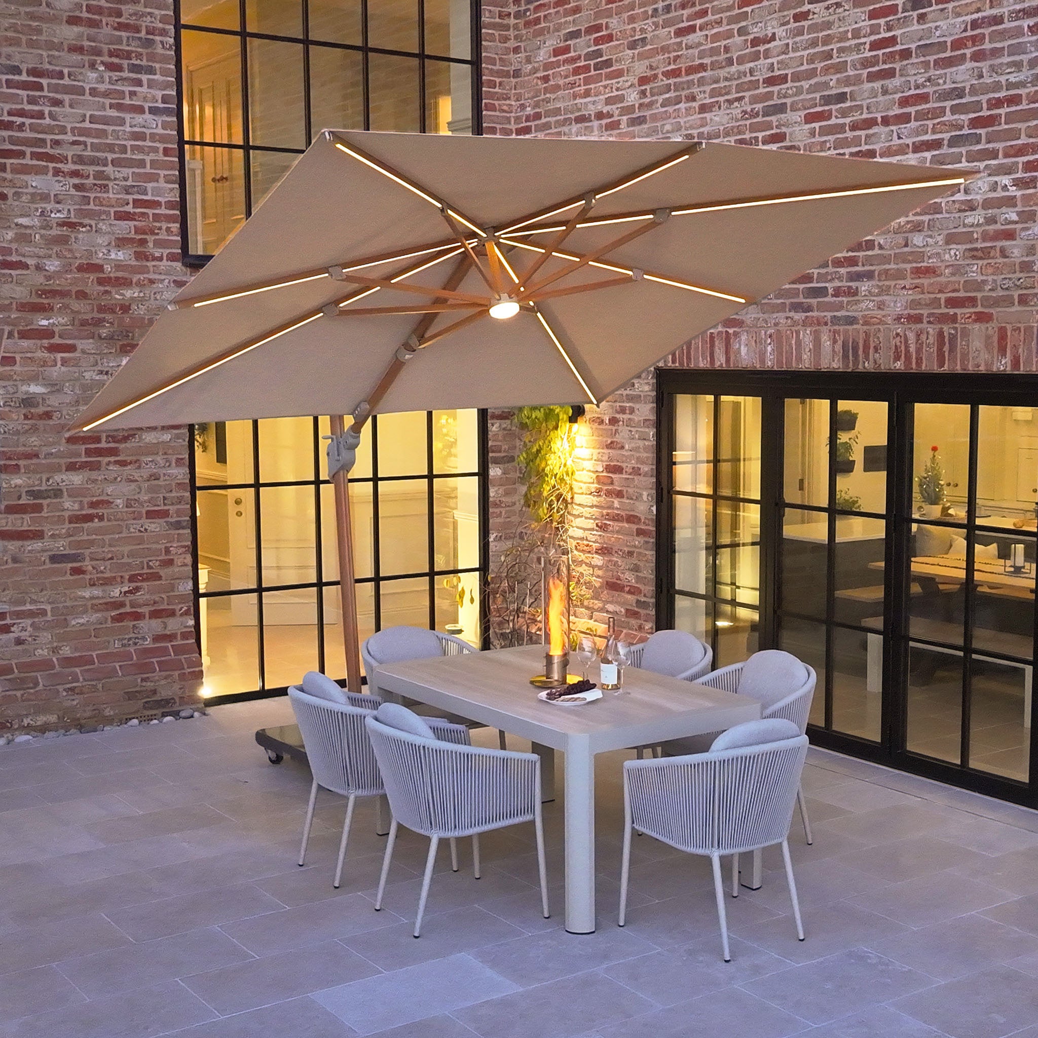 Contemporary outdoor dining area featuring the Volos 3m x 3m Square Cantilever Parasol with LED lighting and a 120kg granite base in Wheat and Wood Effect, complemented by six chairs beside a chic brick wall.