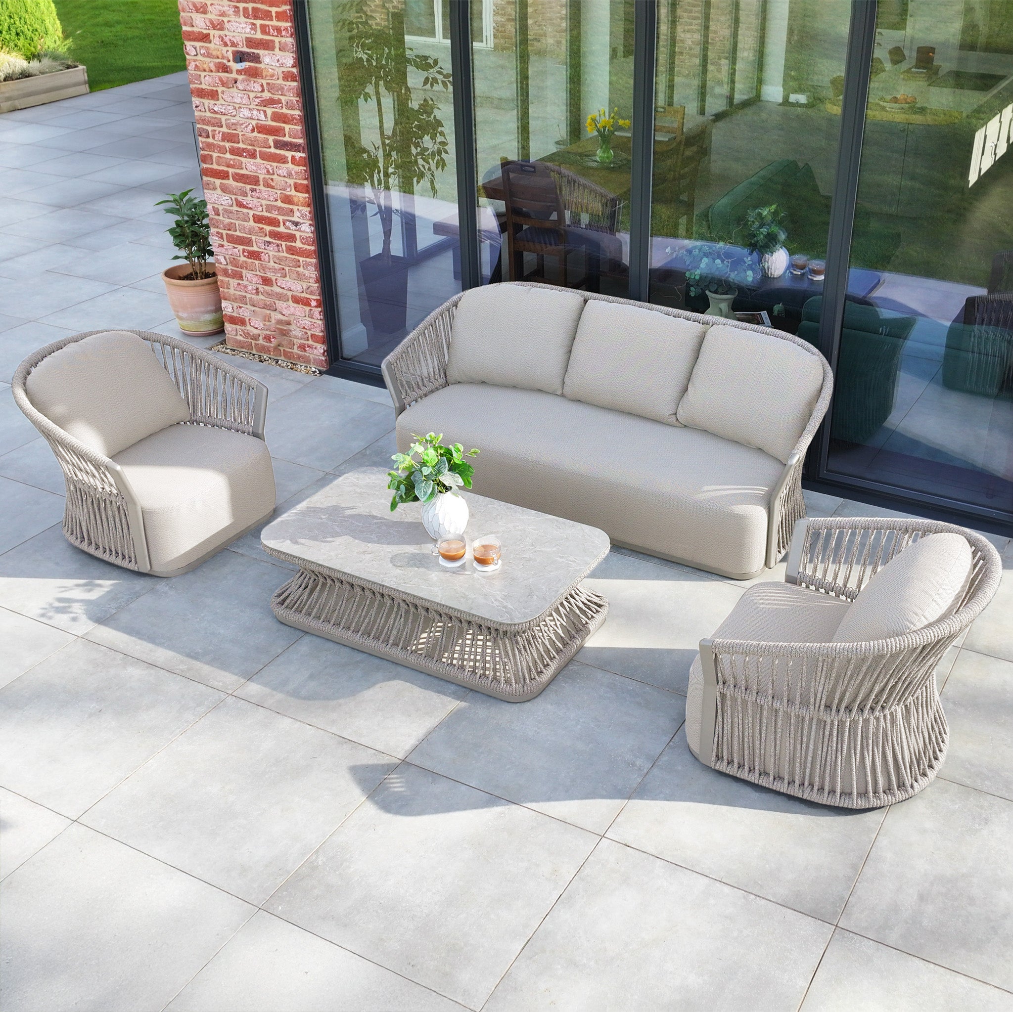 The Pello 3 Seat Sofa Set in Fawn includes an outdoor sofa, two chairs, and a coffee table on a stone patio. Quick Dry Foam cushions offer comfort, while lush plants enhance the tranquility. Solution-dyed olefin fabric ensures durability.