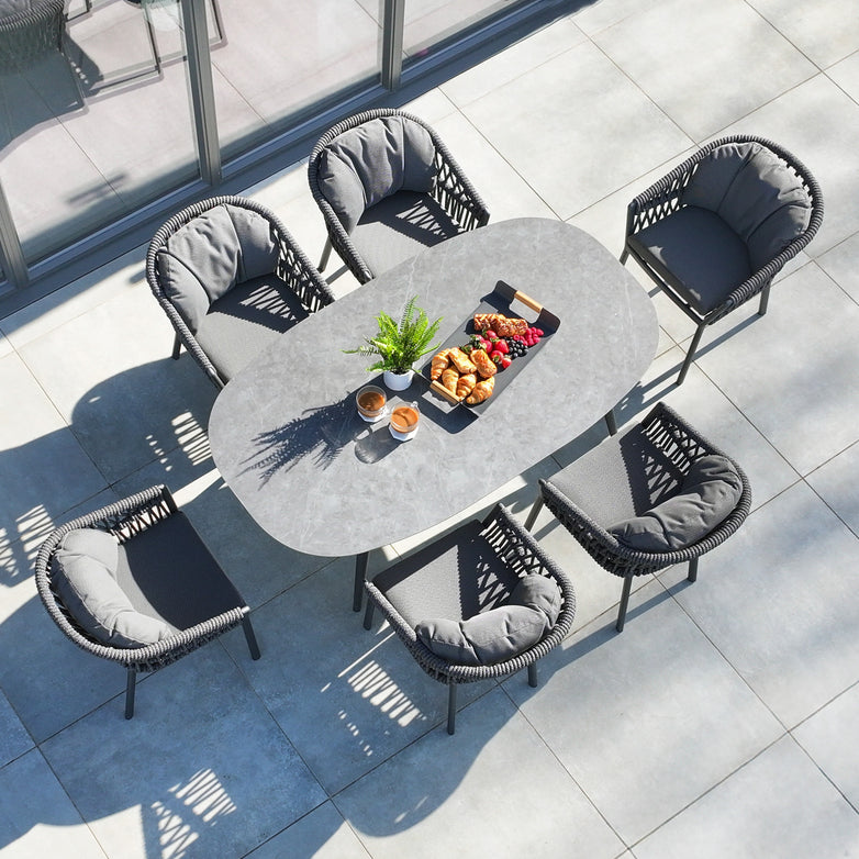 The Pello 6 Seat Oval Dining Set with Ceramic Table in Grey includes a sleek table and six cushioned chairs, complemented by a potted plant centerpiece. This weather-resistant outdoor dining furniture is perfect for long-lasting enjoyment.
