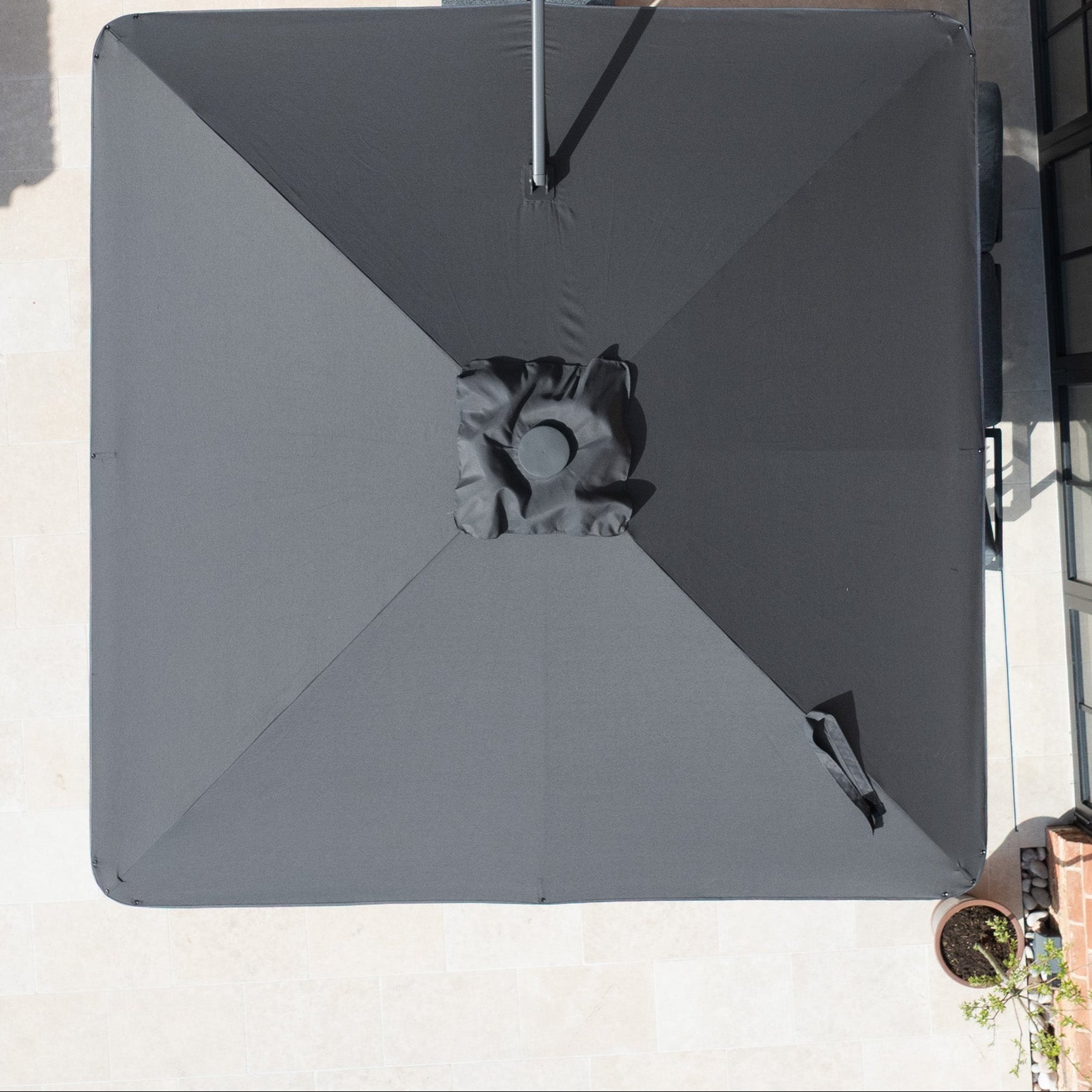 Top-down view of the Nexus T2 Premium 3m Square Cantilever Parasol in Black with 90kg Granite Base on a light stone patio near glass doors and a potted plant.