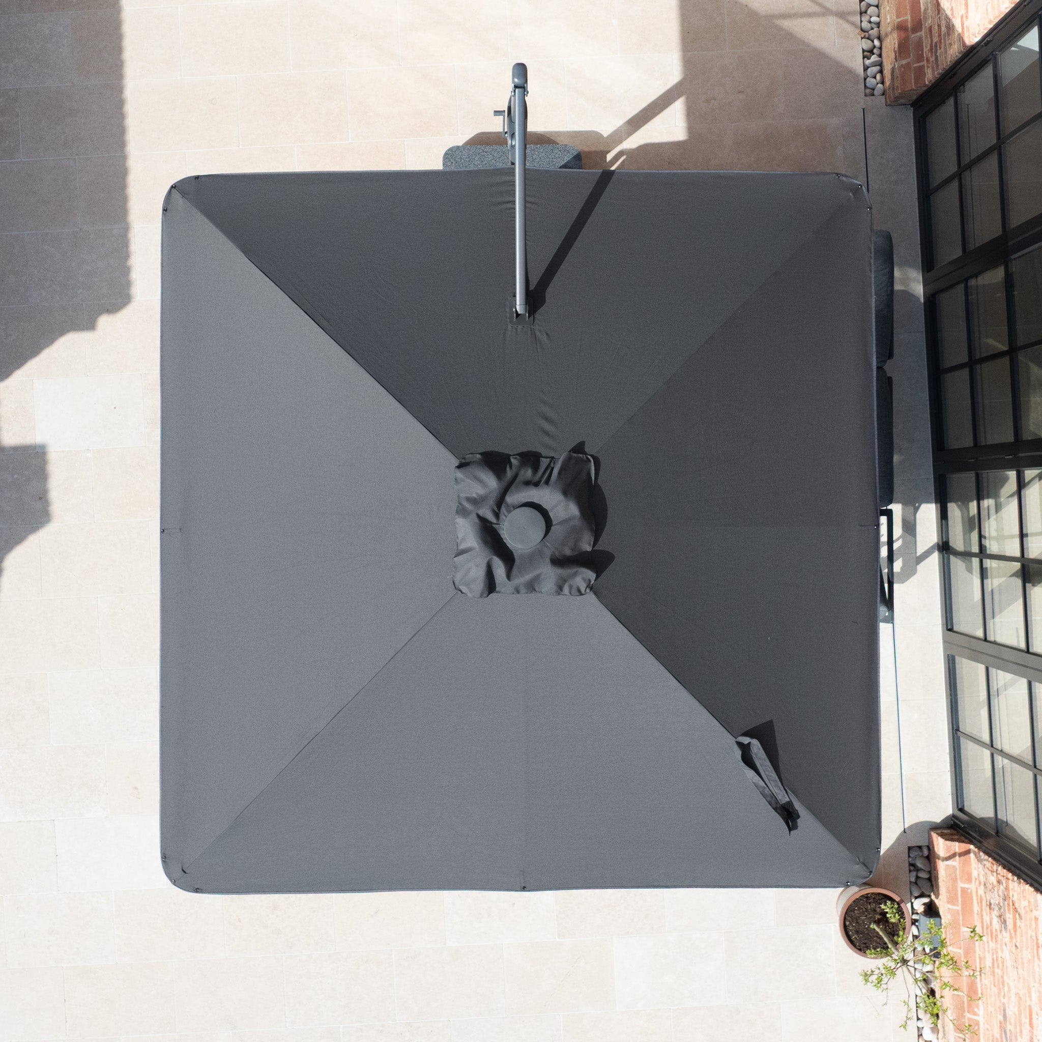 Top-down view of the Nexus T2 Premium 3m Square Cantilever Parasol in Black with 90kg Granite Base on a light stone patio near glass doors and a potted plant.