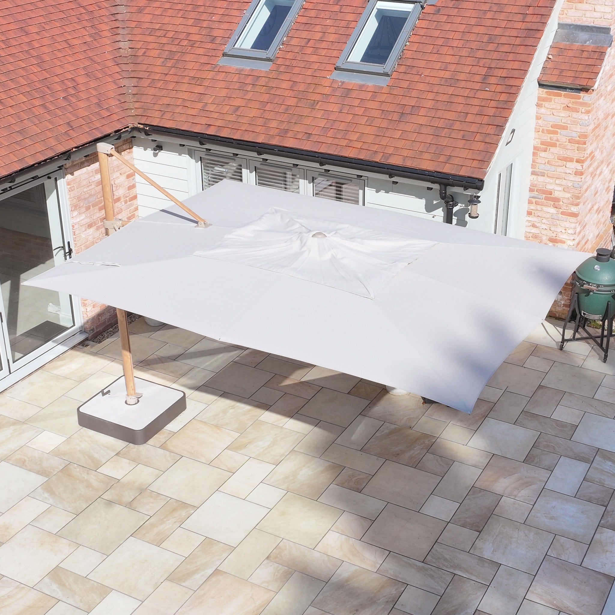 The Finca 4m x 3m Cantilever Parasol in Sand offers essential sun protection on a paved terrace near a brick house with skylights, enhancing your outdoor living experience.