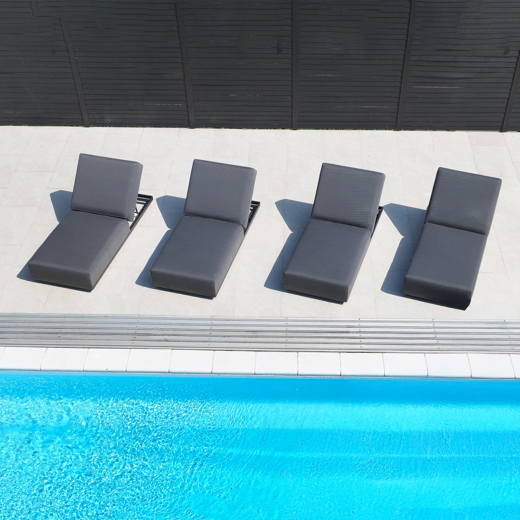 Four empty Luna Outdoor Fabric Deluxe Sun Loungers with Bolster Cushion in Grey are lined up beside a bright blue swimming pool on a sunny day.