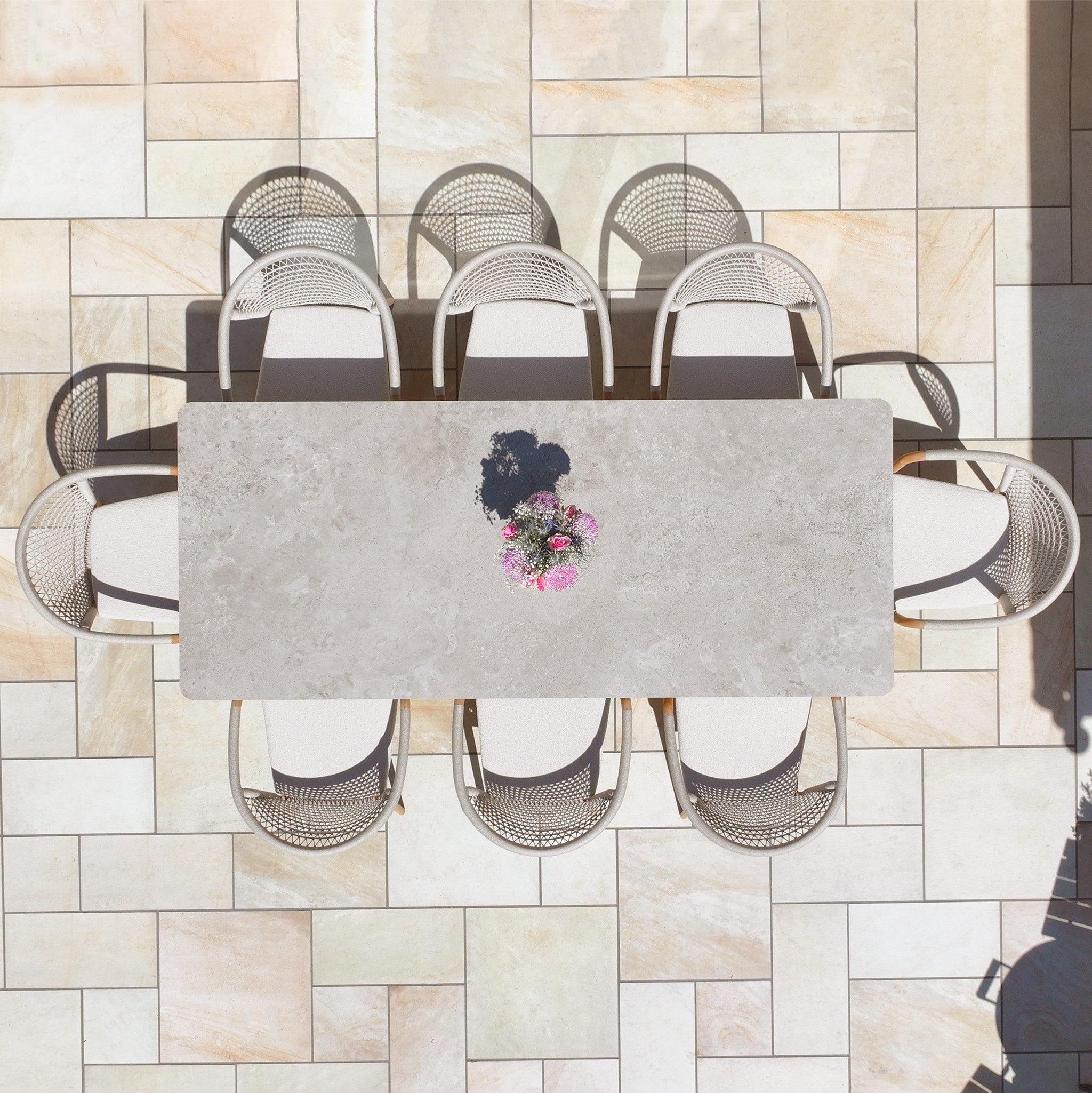 The Sora 8 Seat Rope Dining Set with Ceramic Top Table in Stone features a spacious table and eight stylish chairs, elegantly accented with a small bouquet of pink flowers at the center.