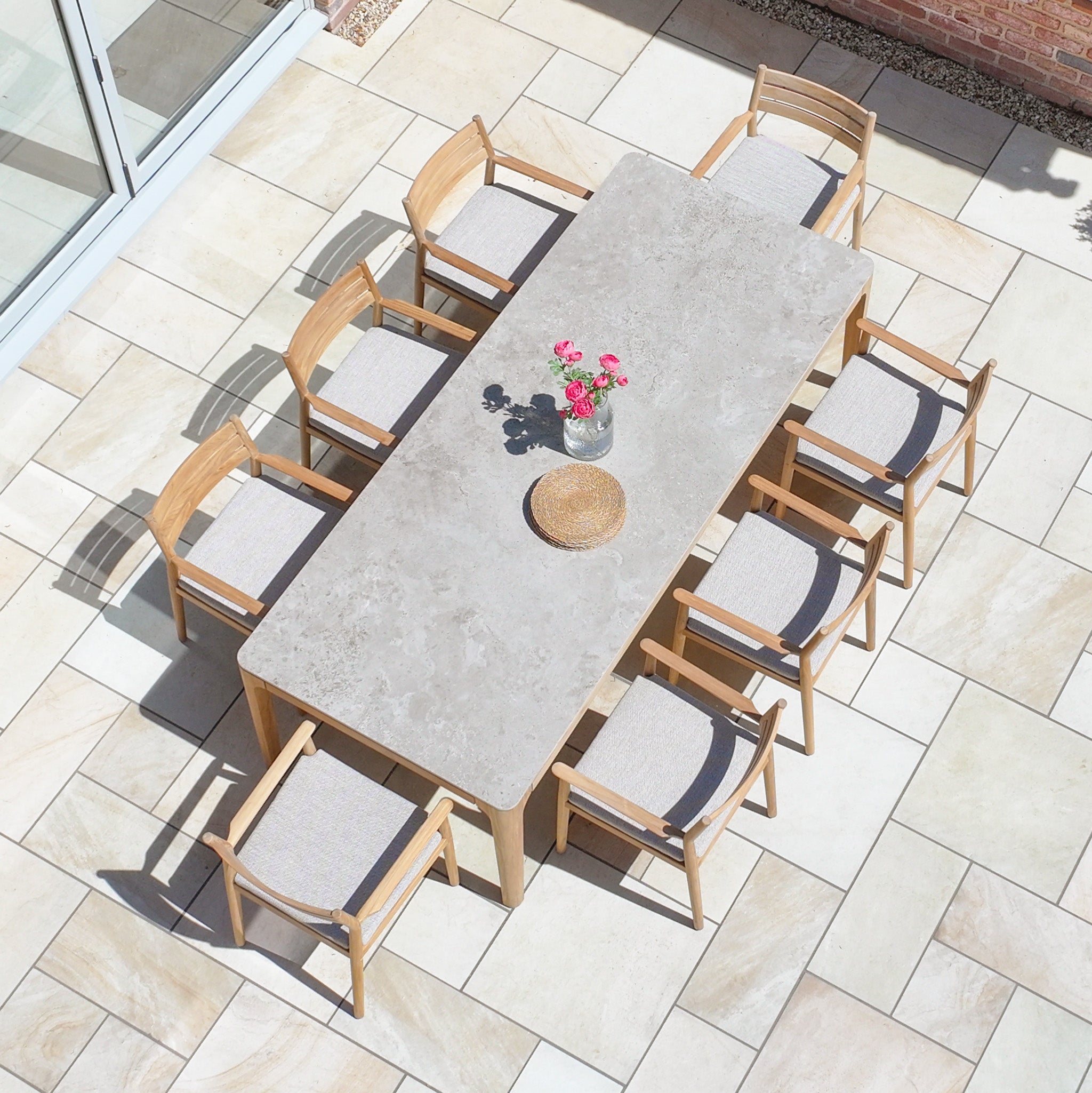 Siena 8 Seat Dining Set with Ceramic Top Table in Fawn, shown with eight chairs, a flower vase, and a woven tray on a tiled patio.