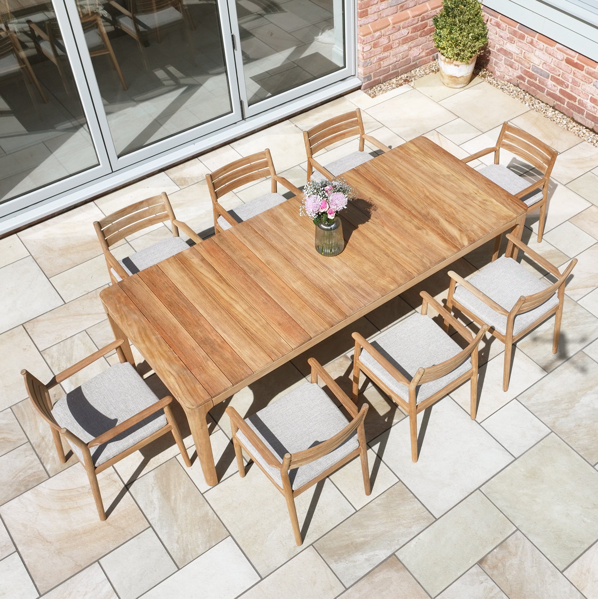 The Siena 8 Seat Dining Set with Teak Table in Fawn, arranged on a tiled patio by glass doors and a potted plant, is ideal outdoor furniture for gatherings.