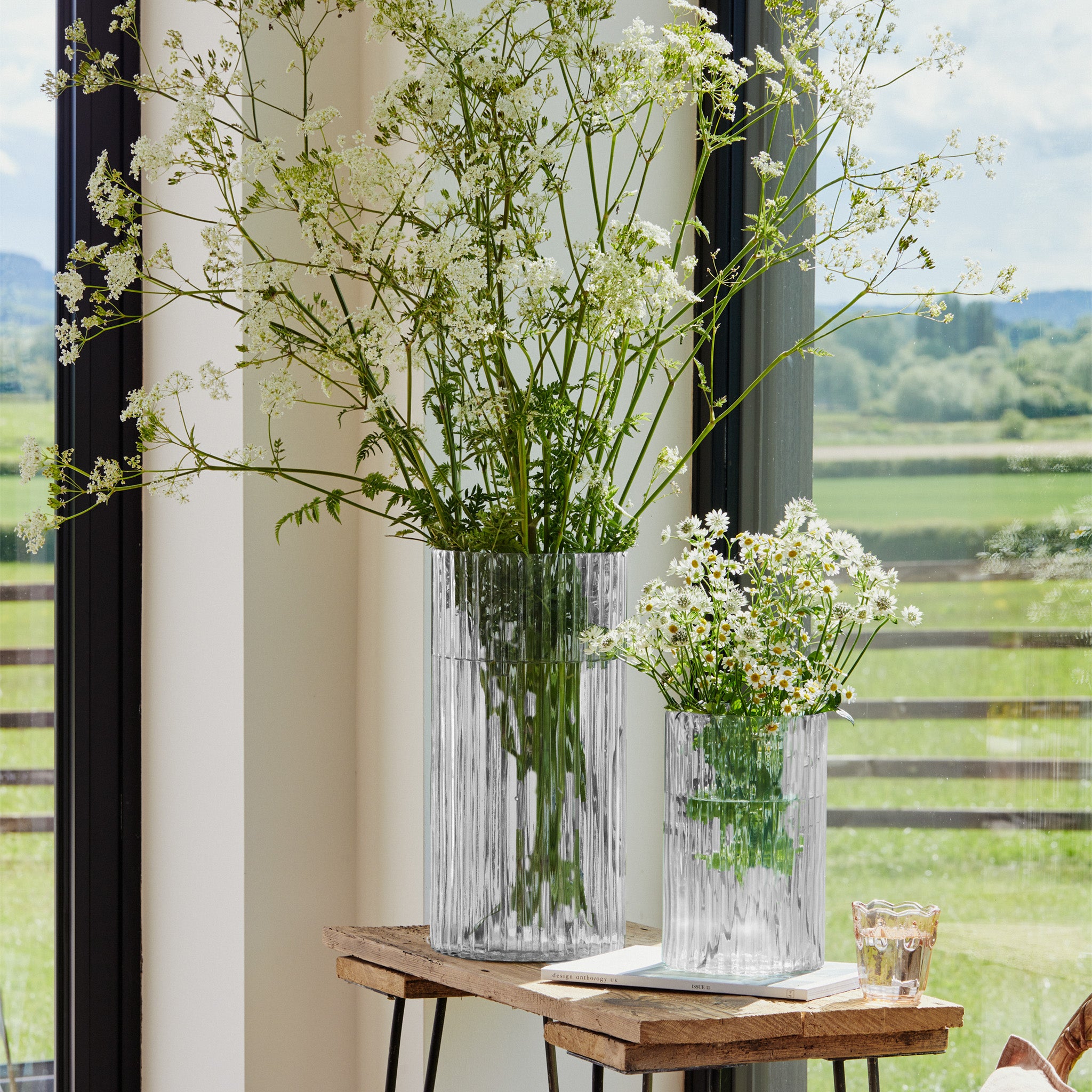 Two Ivyline Daphne Ribbed Vases in Clear, filled with wildflowers, sit on a wooden table. They add a touch of sustainable interior décor to the scene, framed by large windows overlooking the scenic countryside.