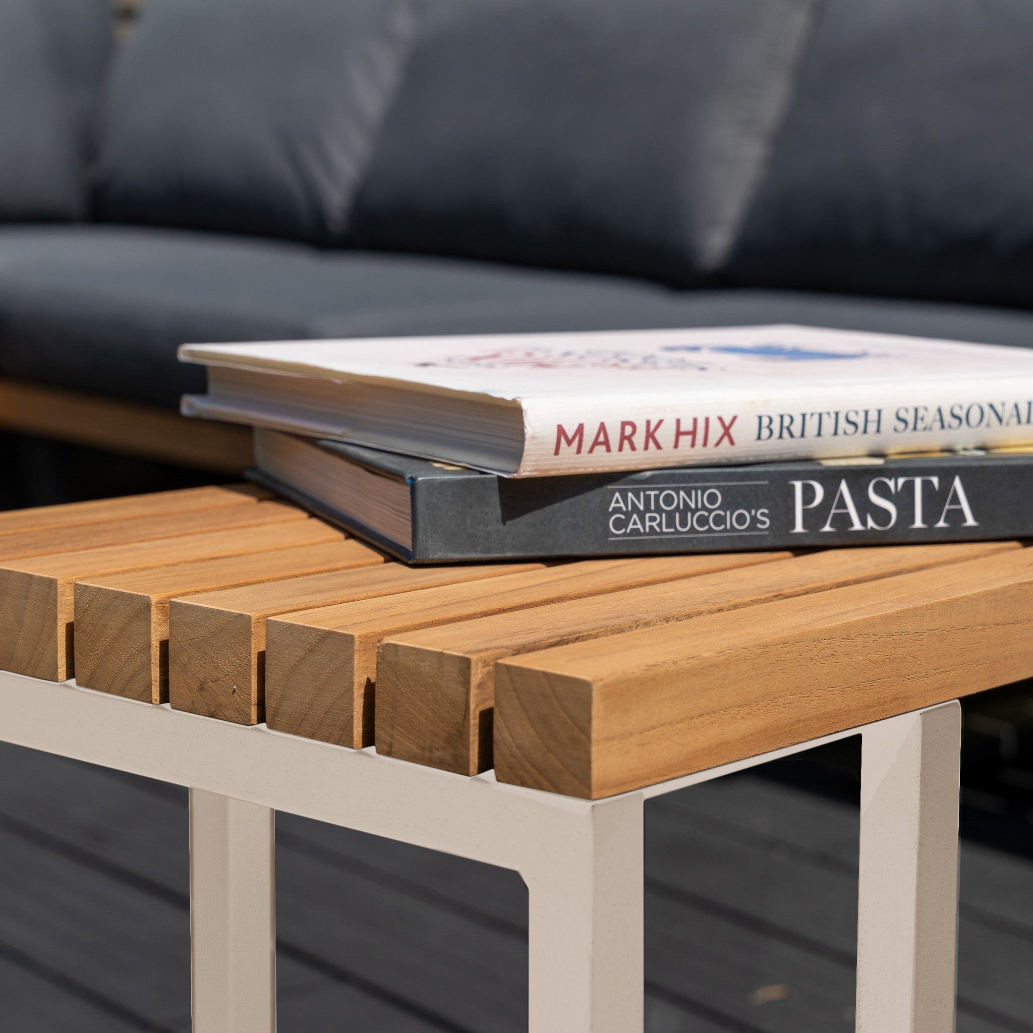 Three books are stacked on the Cube Side Table with Teak Top in Latte, displayed outdoors with a sofa in the background.