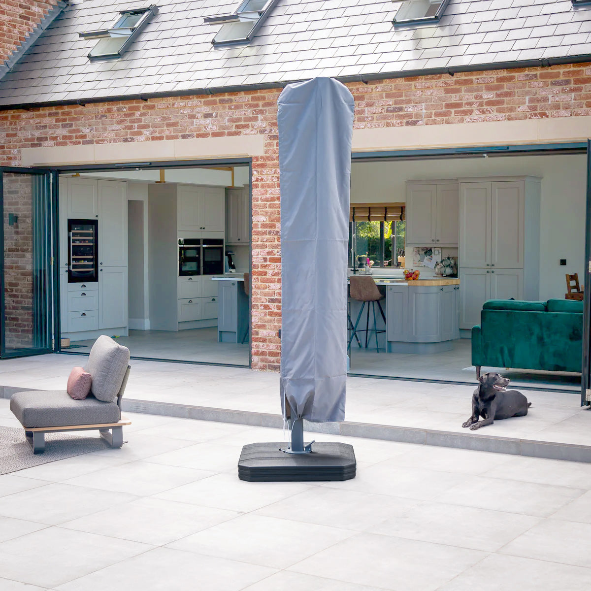 The patio is sheltered by the Ares 3.5m Round Cantilever Parasol with a Solar-powered LED-lit UV50+ canopy in Beige, while modern furniture complements the open kitchen. Nearby, a dog relaxes in this spacious outdoor haven.