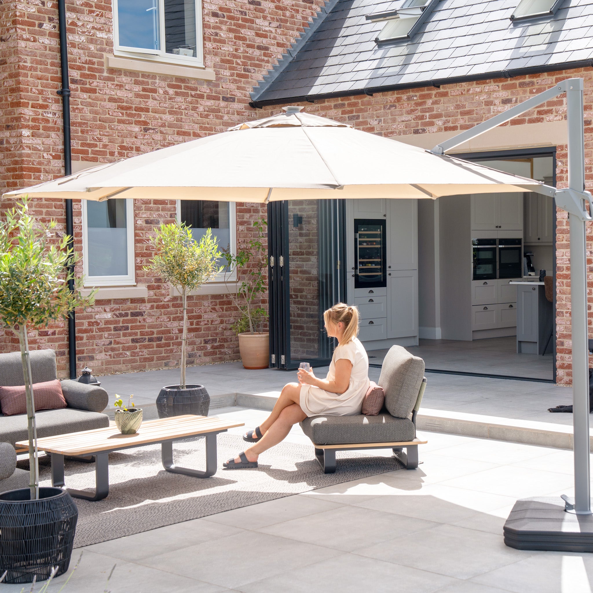 A woman relaxes beneath the Ares 3.5m Round Cantilever Parasol with solar-powered LED lights in beige on a modern backyard patio with sleek furniture and vibrant plants.