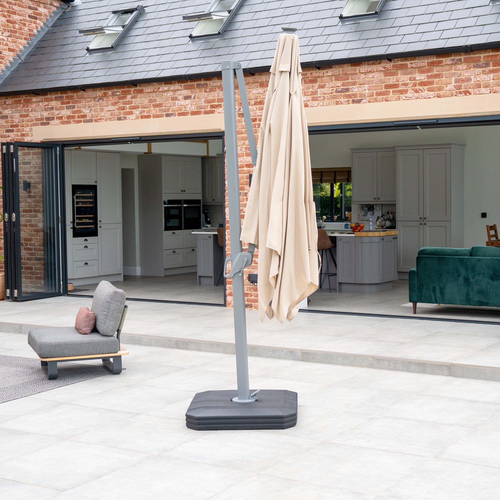 Outdoor patio showcasing a modern lounge chair and the Ares 3m Square Cantilever Parasol with Solar Powered LED Lights in Beige, offering an open view to a spacious kitchen and living area.