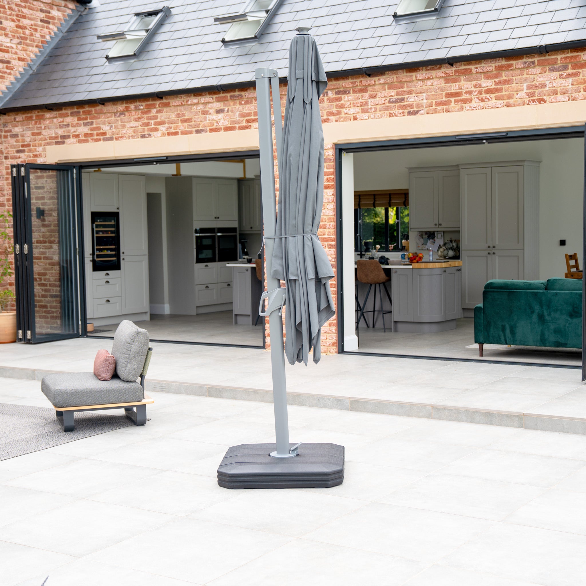 On a terrace with a modern brick house, open doors, and outdoor seating, the closed Ares 3m Square Cantilever Parasol in Charcoal offers ideal daytime lounging. Its UV50+ resistance ensures sun protection.
