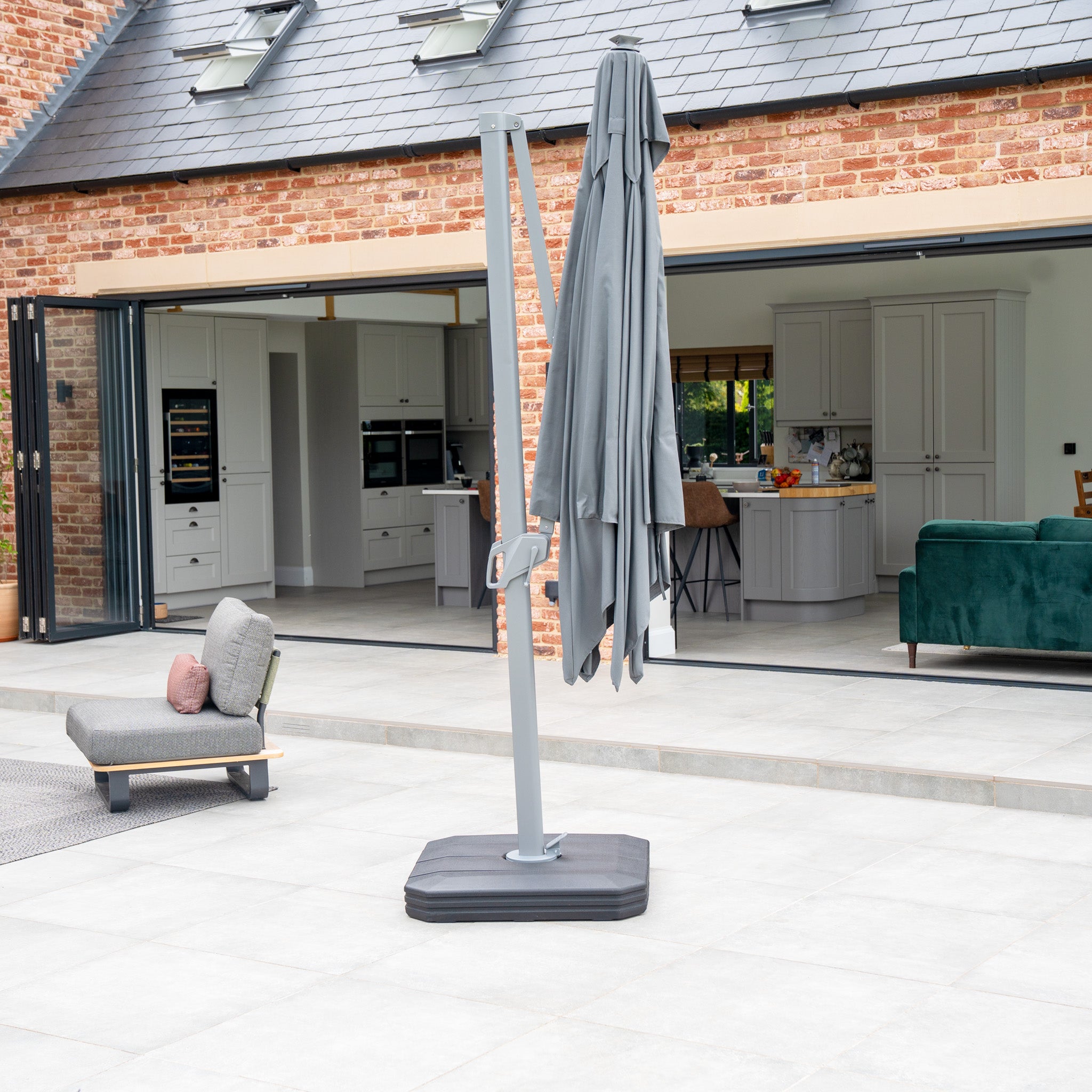 Patio featuring a closed Ares 3m Square Cantilever Parasol with Solar powered LED Lights in Charcoal, lounge chair, and open sliding doors leading to a modern kitchen.