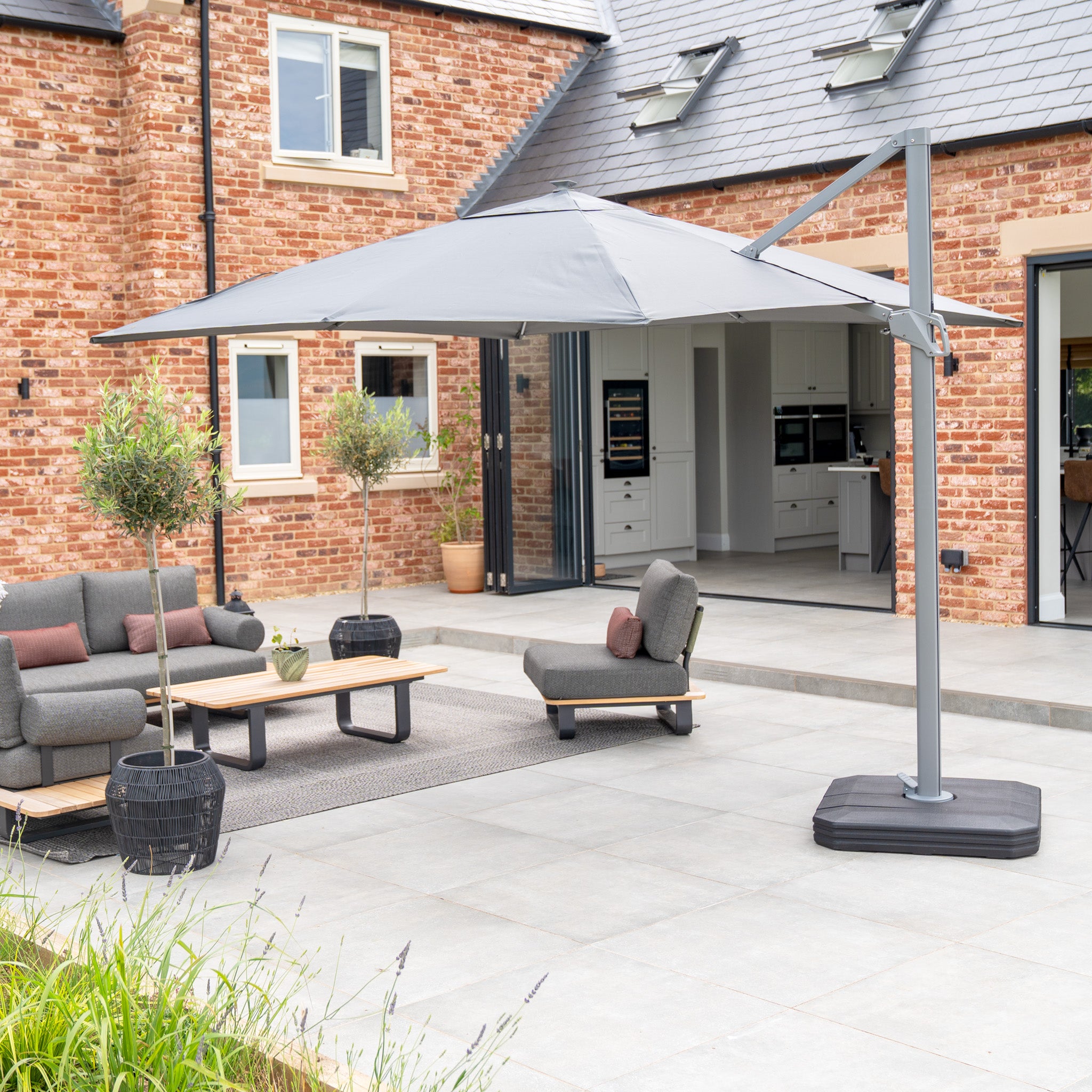 A patio with grey furniture and a 3m Ares cantilever parasol, featuring a charcoal UV50+ canopy and solar-powered LED lights, sits beside a brick house with modern windows and doors.