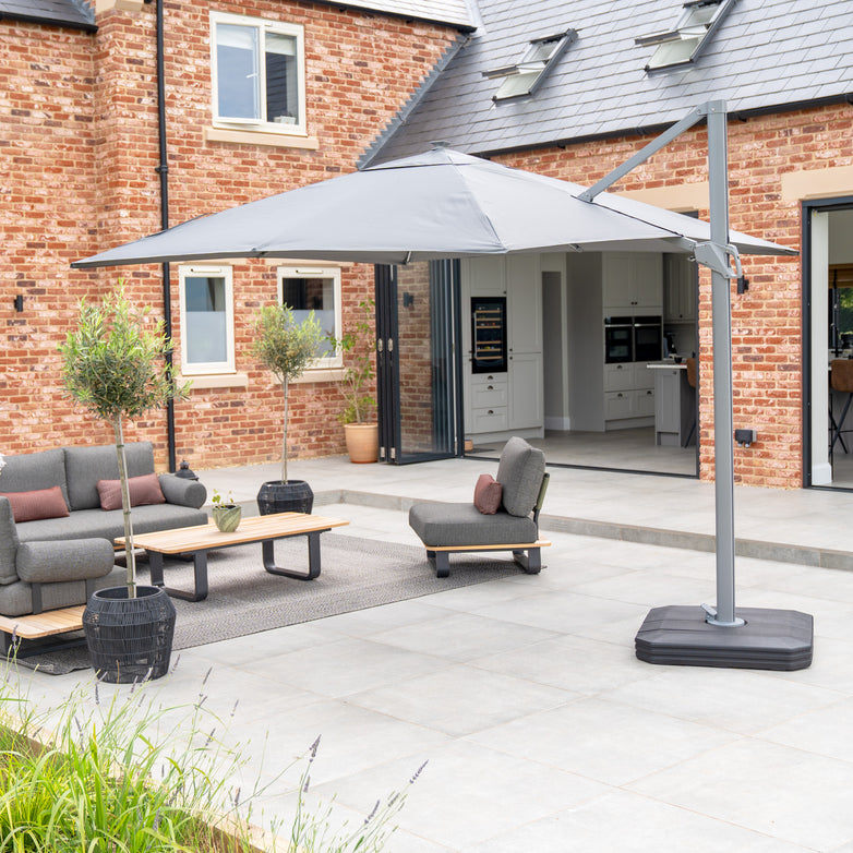 A patio with grey furniture and a 3m Ares cantilever parasol, featuring a charcoal UV50+ canopy and solar-powered LED lights, sits beside a brick house with modern windows and doors.