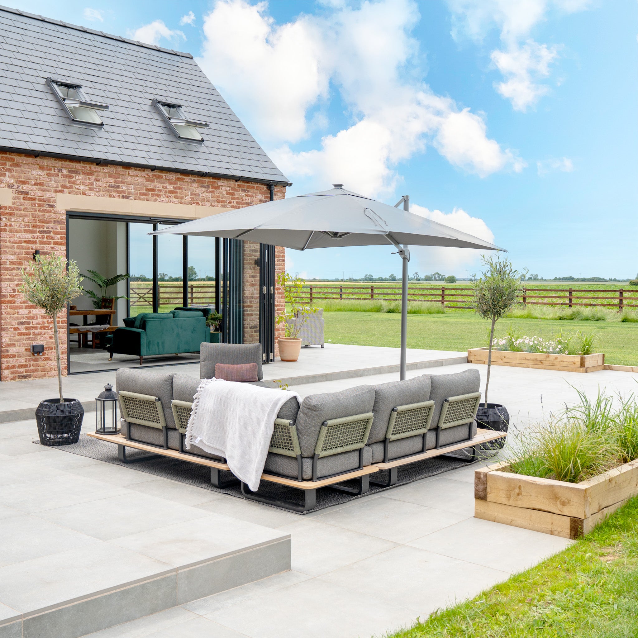 The modern patio has gray seating under an Ares 3m Square Cantilever Parasol with a UV50+ canopy in Charcoal, featuring solar-powered LED lights. Open doors connect to the house as clouds pass over the lush green lawn in the background.