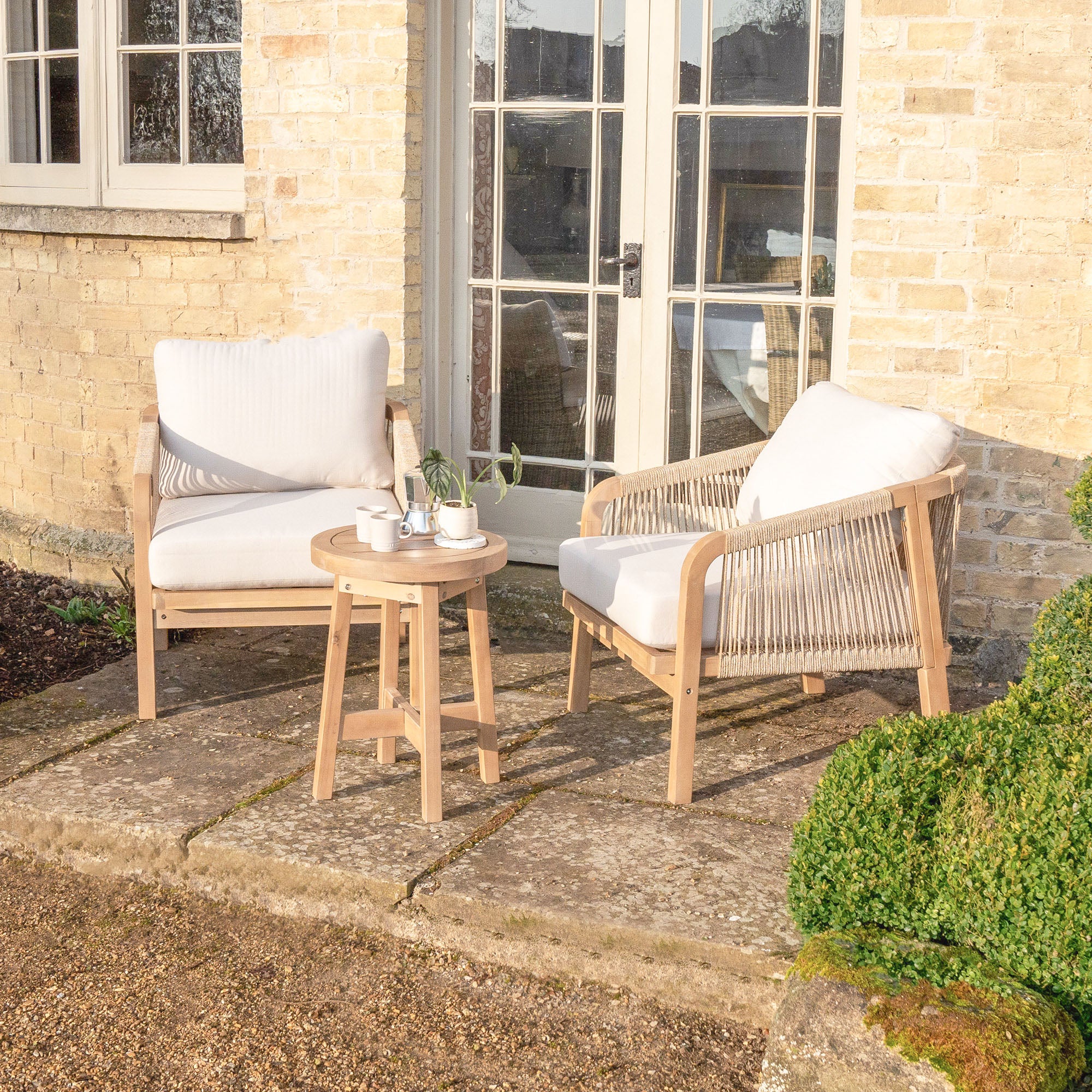 The Quay Lounge Set in Linen, featuring two cushioned chairs with rope weave backs and a round acacia wood table, creates an inviting outdoor lounging area on this brick house's stone patio.