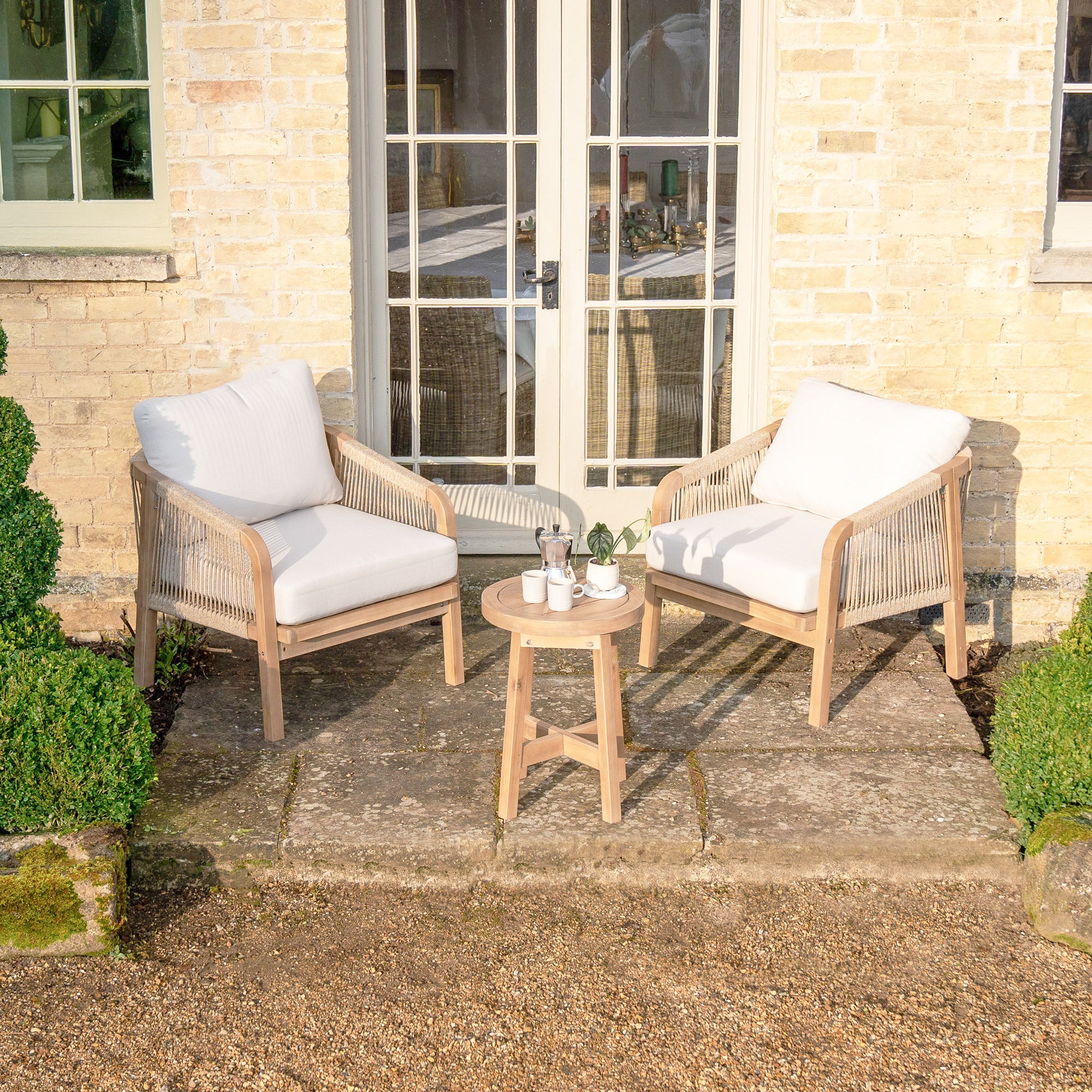 The Quay Lounge Set in Linen, featuring two cushioned rope-weave chairs and a compact acacia wood table, creates a cozy outdoor lounging area on the stone patio.