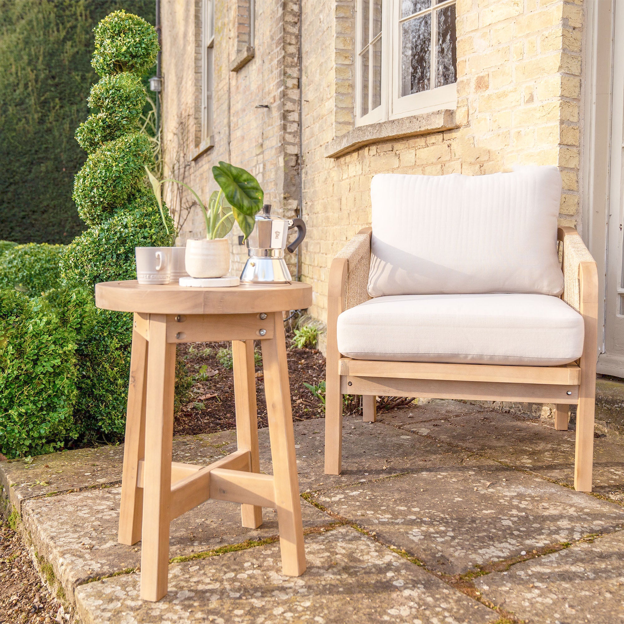 The Quay Lounge Set In Linen is arranged on a stone patio outside a brick house, complete with a coffee set and green topiary nearby—creating an inviting outdoor lounging area.