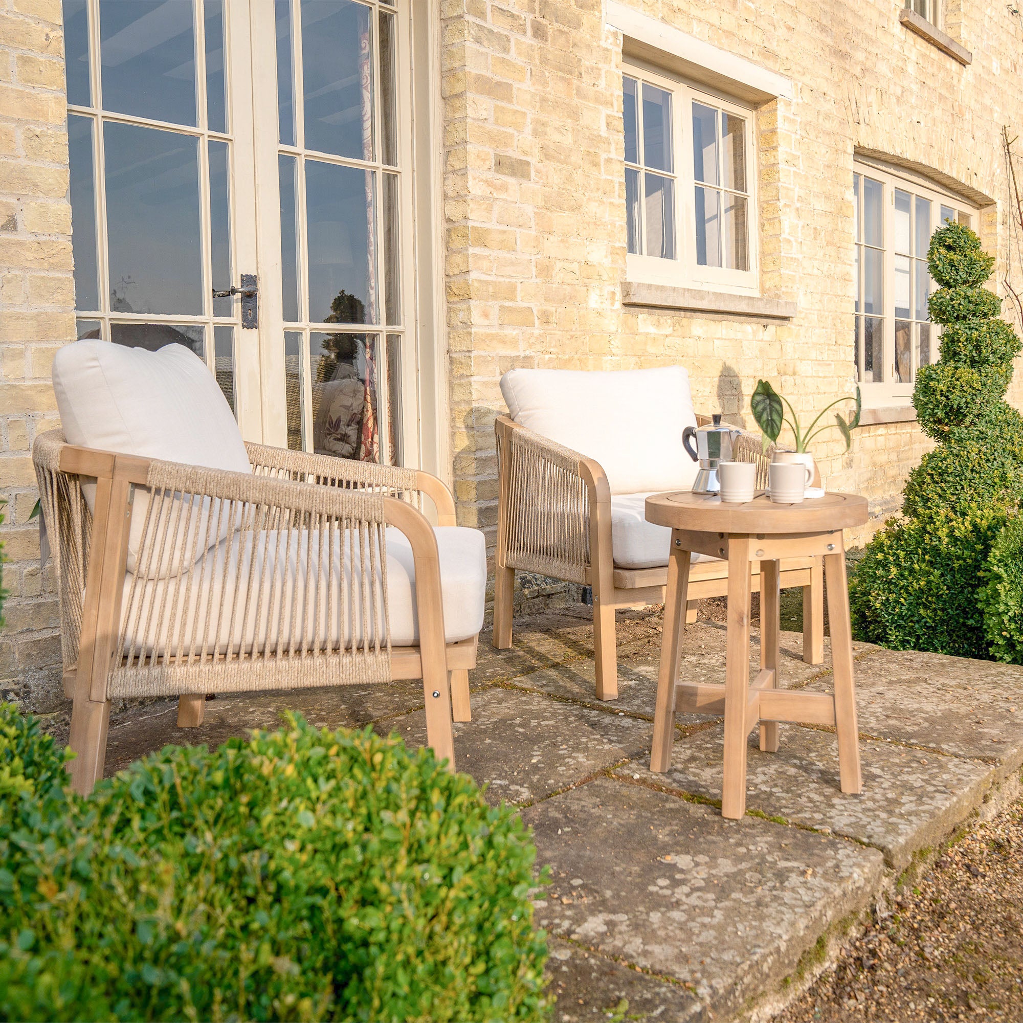 The Quay Lounge Set in Linen features two wooden chairs with rope weave backs and white cushions beside a small table, creating an inviting outdoor lounging area on a sunny stone patio outside a brick house.