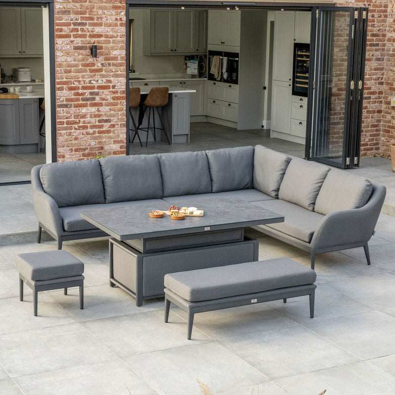 Modern outdoor patio featuring the Luna Outdoor Fabric Rectangular Corner Dining Set with Rising Table in Grey (Right Hand), including a grey sectional sofa, matching stools, and a rectangular table in front of an open-plan kitchen.